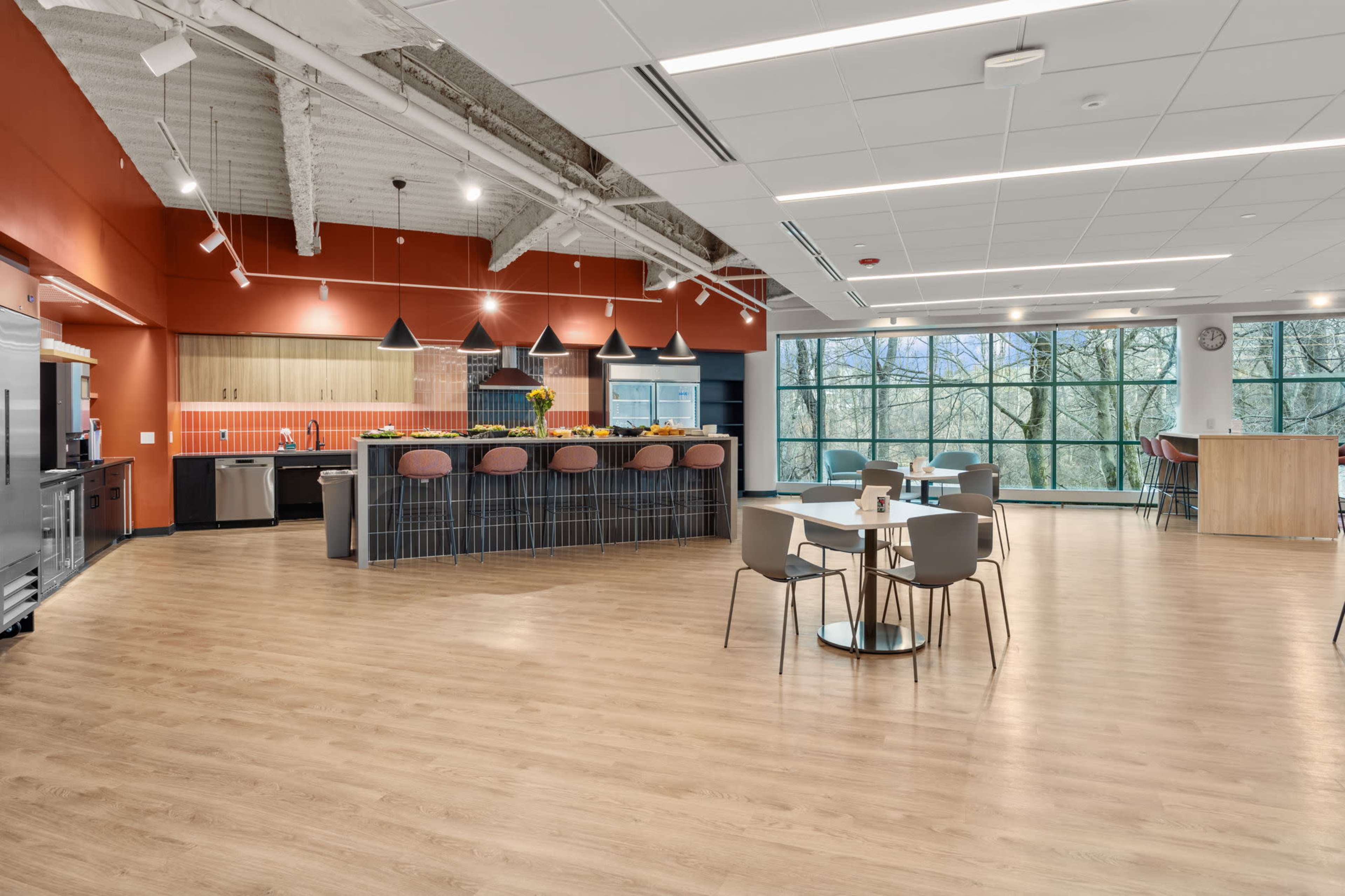 A modern kitchen and dining area with a large window overlooking a natural landscape, featuring a central island with bar seating and various kitchen appliances.
