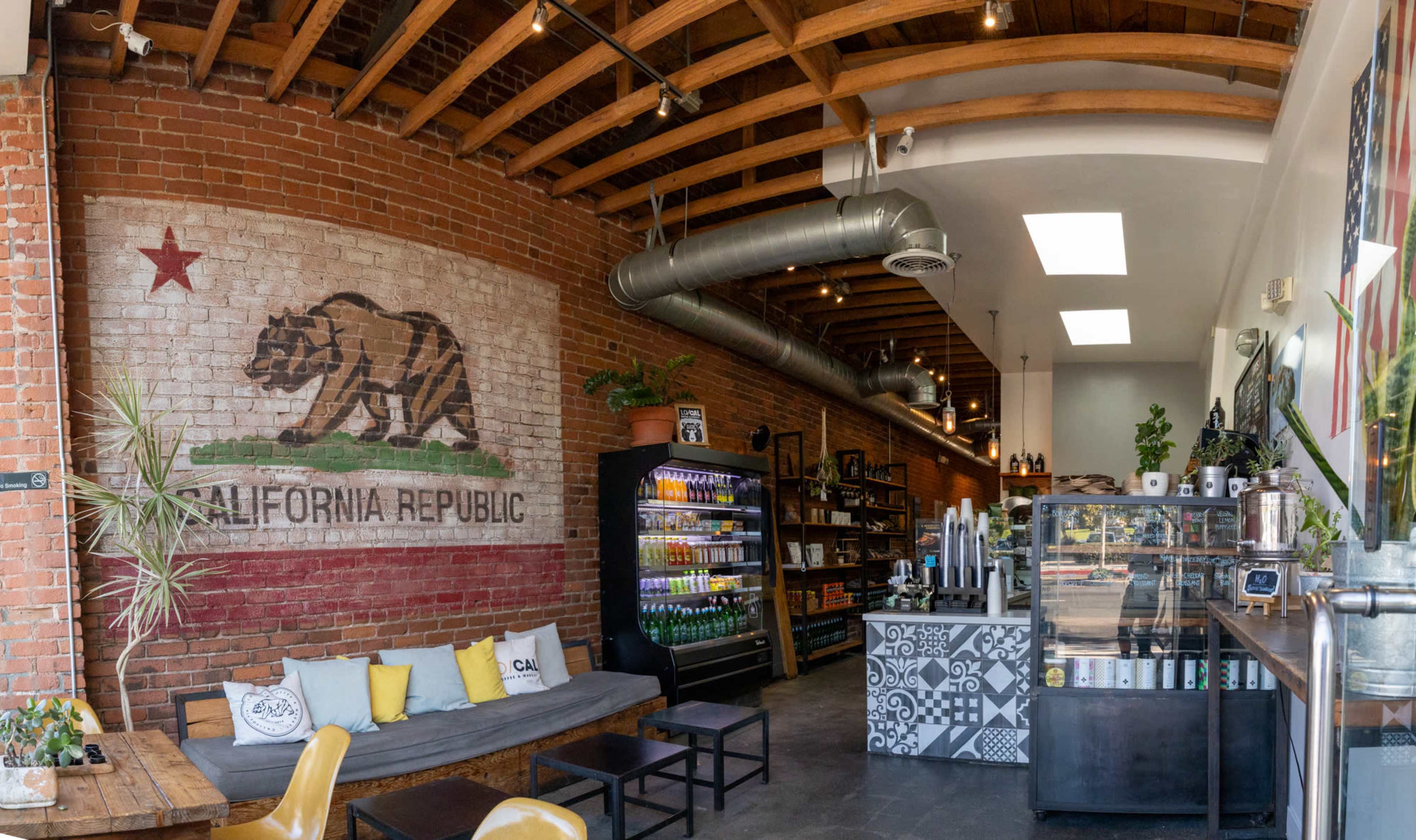 A café interior featuring a large mural of the California flag on a brick wall, a seating area with a long sofa, a refrigerator stocked with beverages, and a counter with coffee equipment.