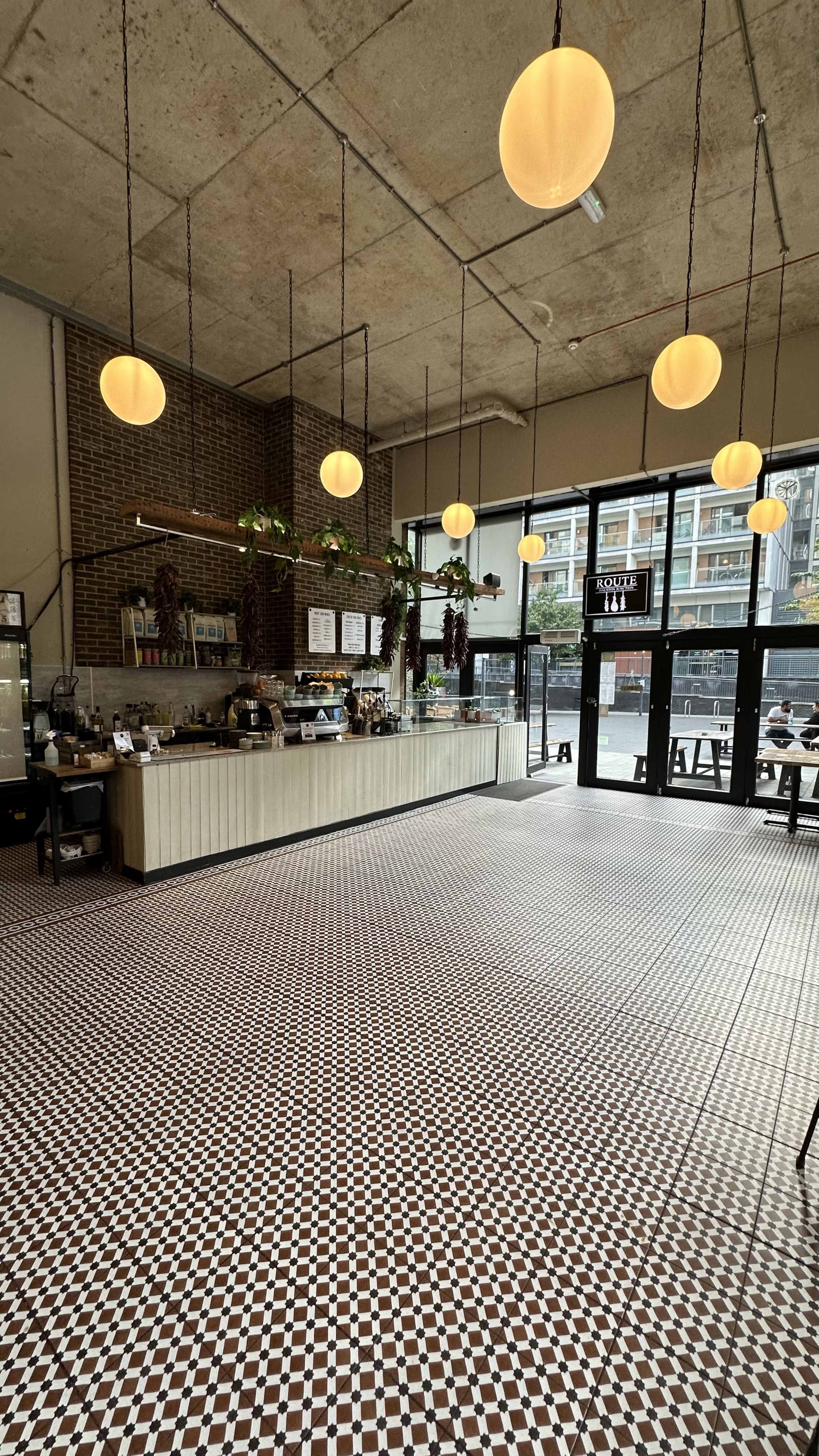 The image shows a spacious cafe with a patterned tile floor, a long counter stocked with food and drinks, and large windows allowing natural light to enter.