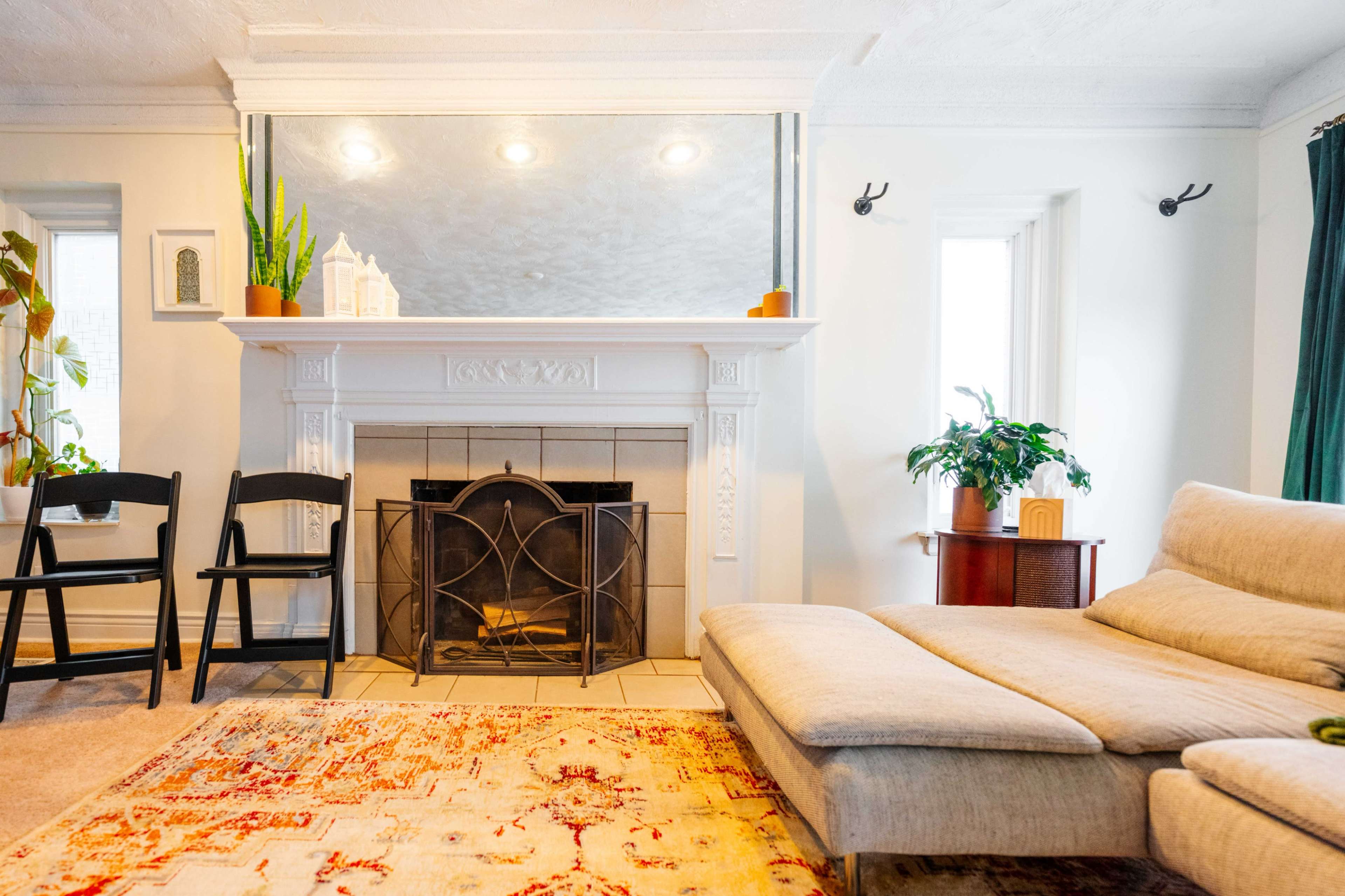 The image shows a living room with a fireplace, a sectional sofa, a patterned rug, and several plants.