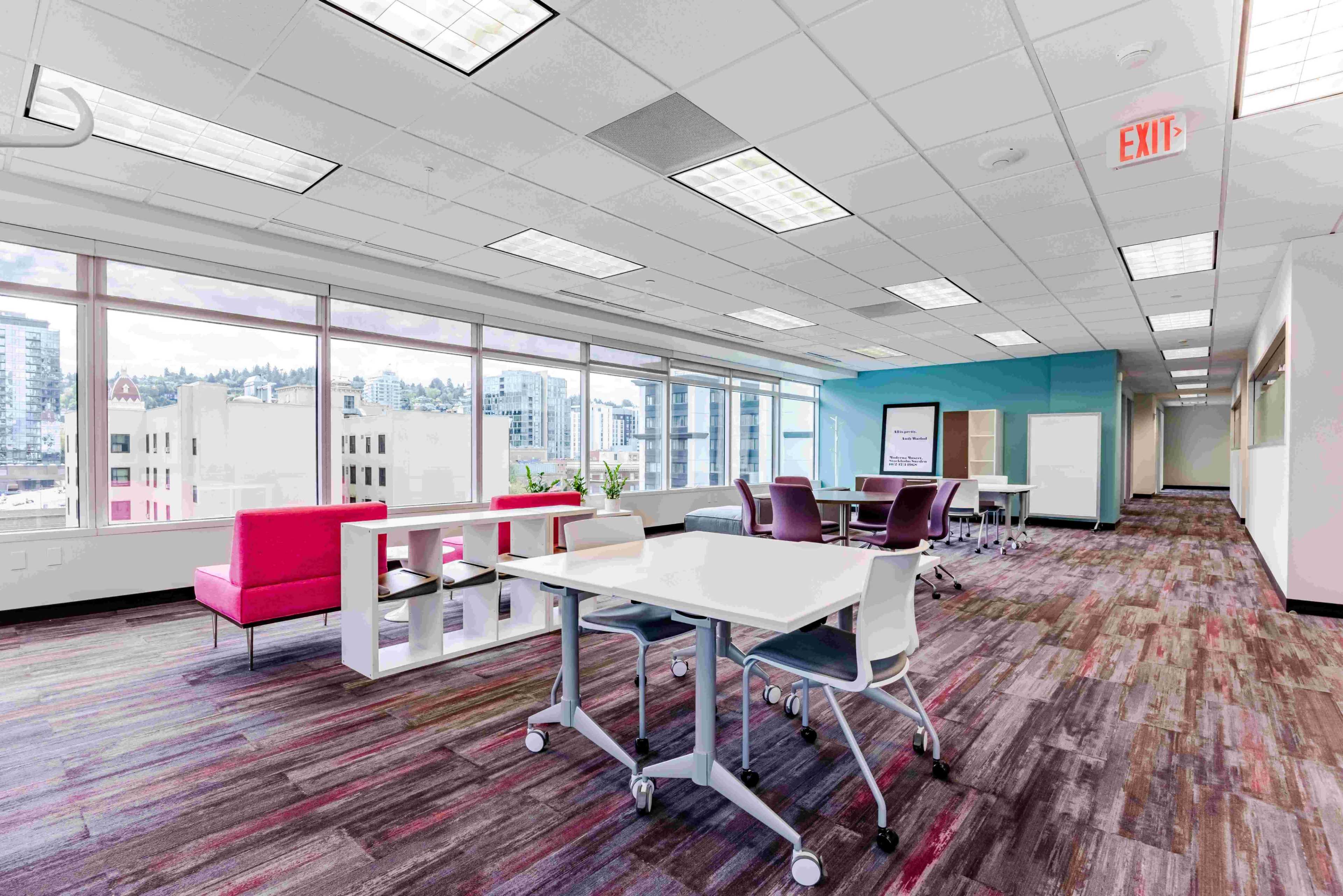The image shows a modern office space with large windows, colorful carpeting, and various workstations arranged throughout the room.