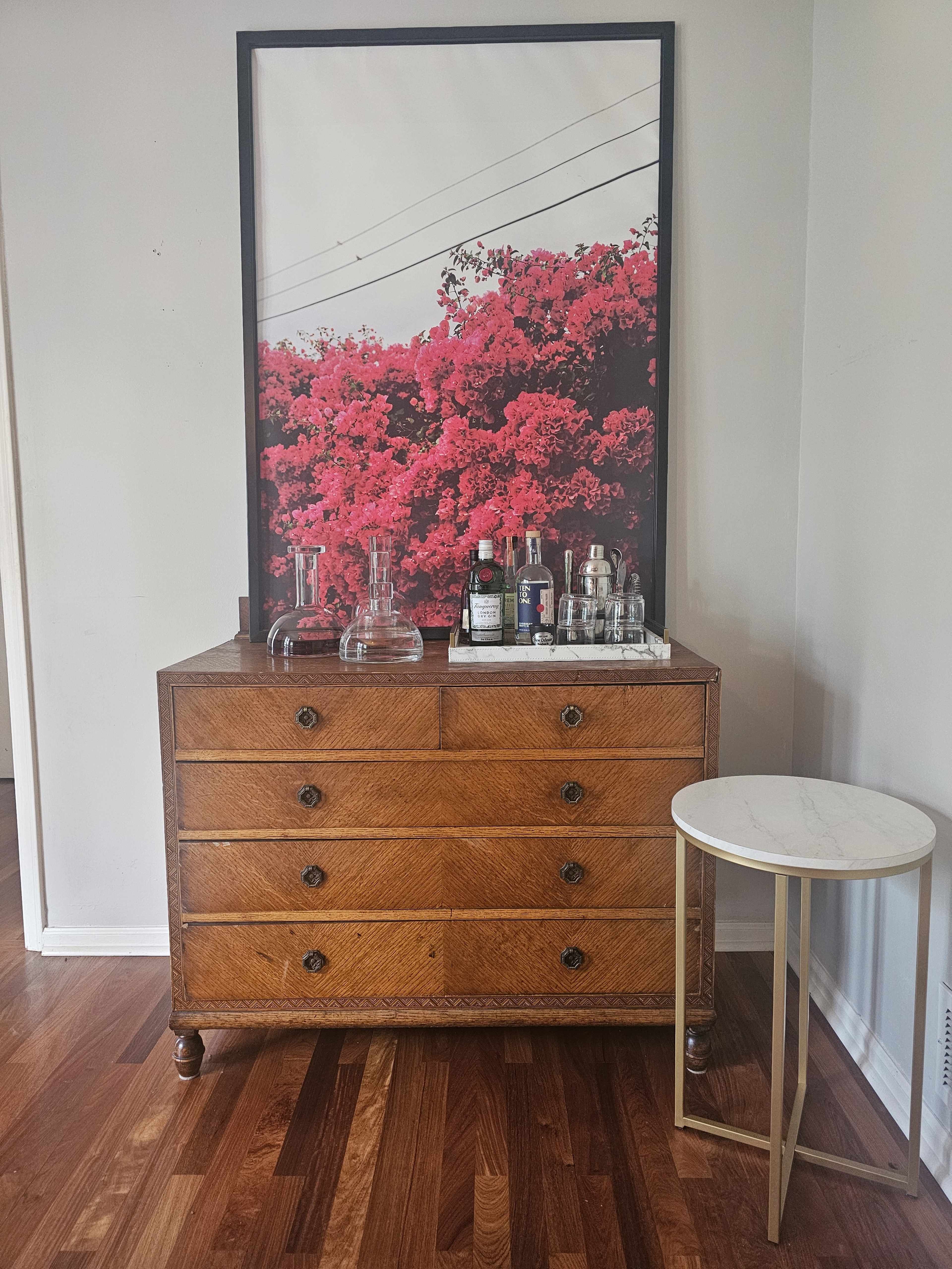 A wooden dresser with six drawers is placed next to a small round table, featuring a framed vibrant floral photo and several glass decanters and bottles on top.