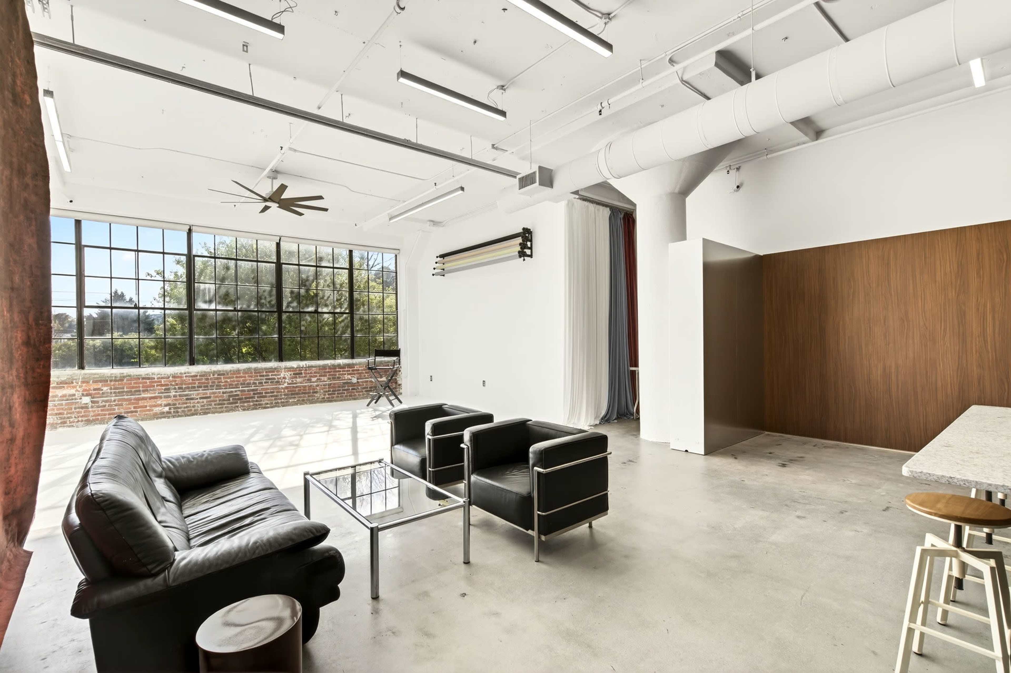 The image shows a spacious, modern interior with a large window, industrial-style lighting, and minimalist furniture, including a black sofa and two armchairs.