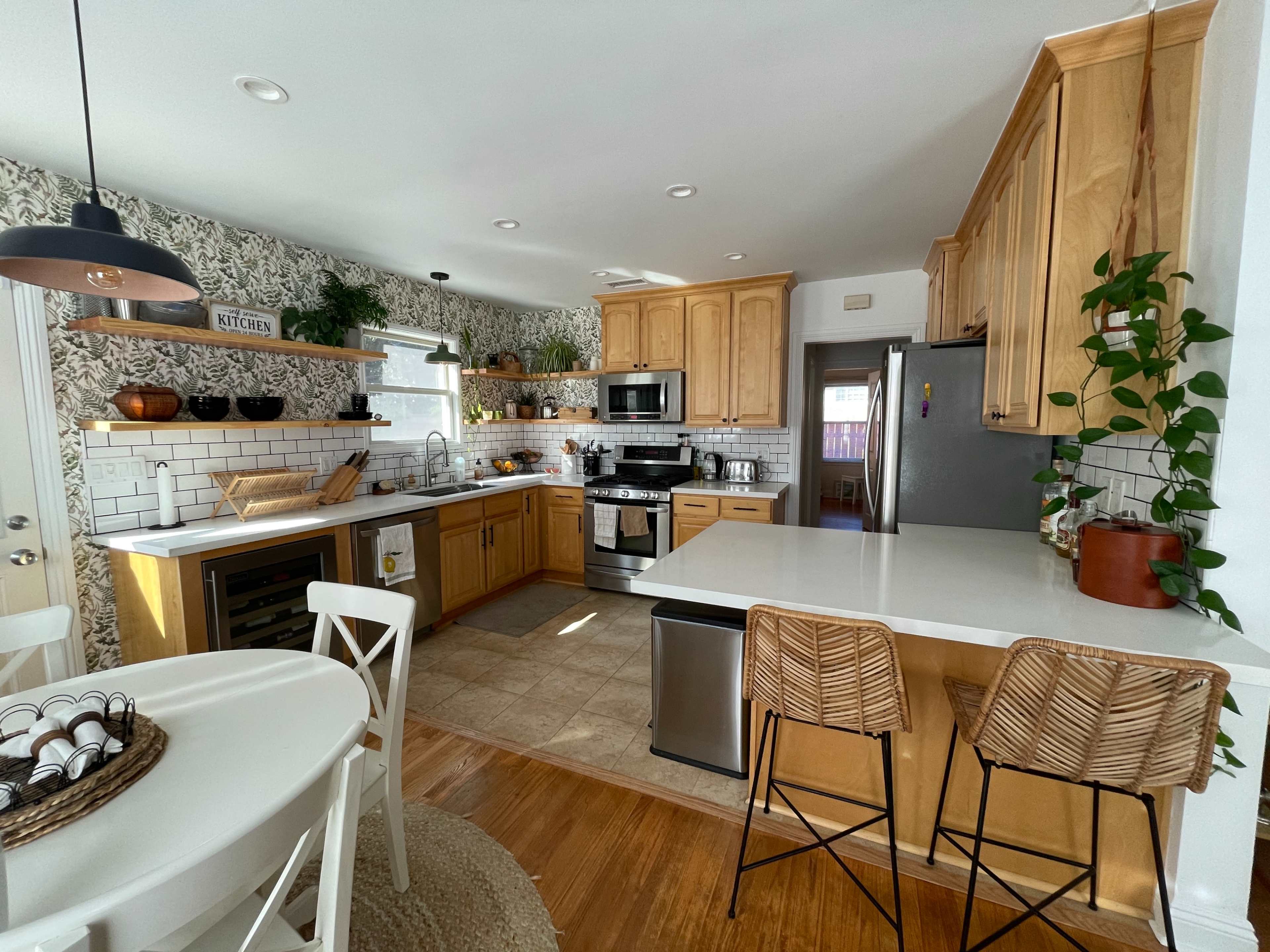 The image shows a modern kitchen with wooden cabinets, stainless steel appliances, a white countertop, and a dining area with a round table and bar stools.