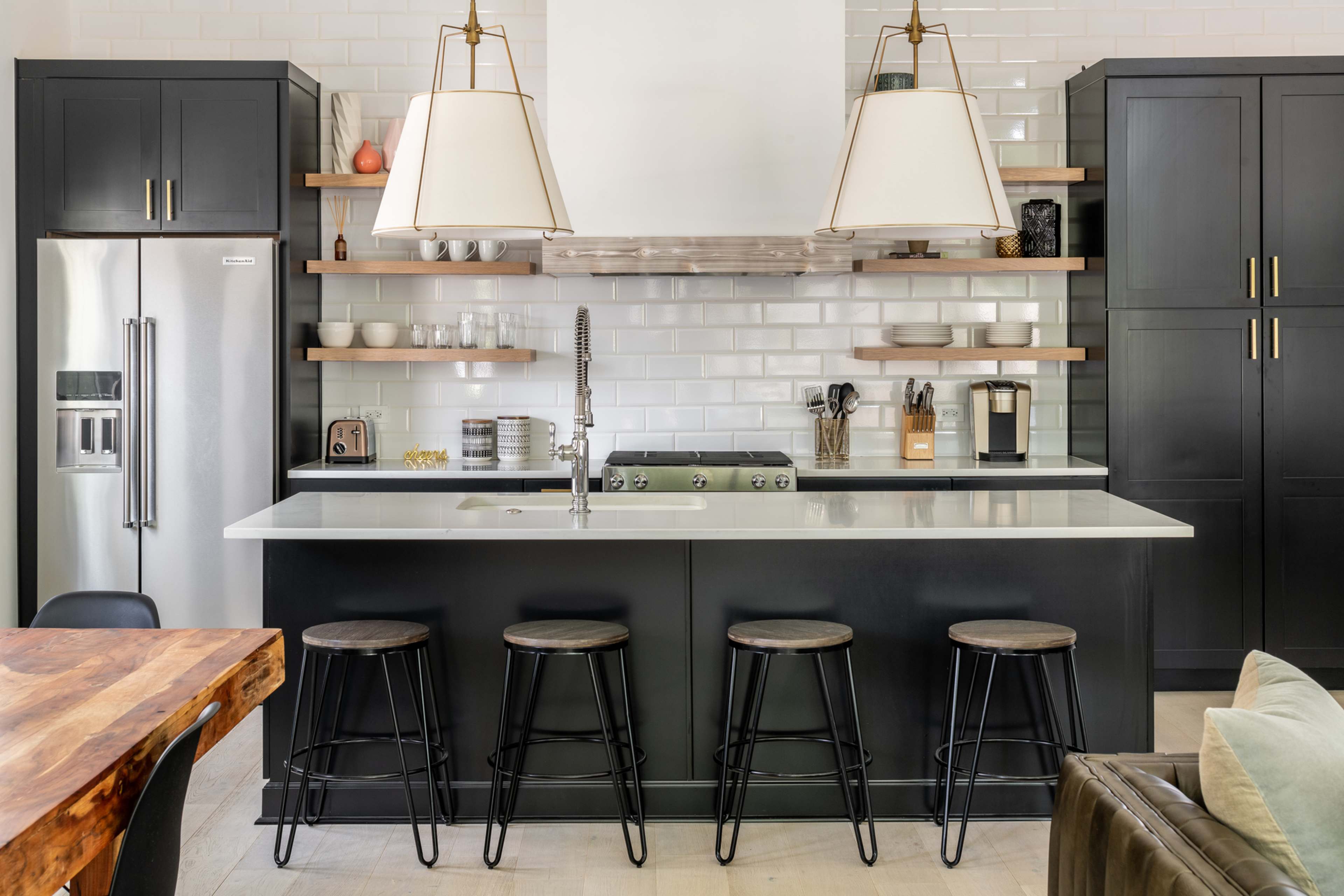 A modern kitchen with black cabinetry, a white countertop, and four metal stools at the island.