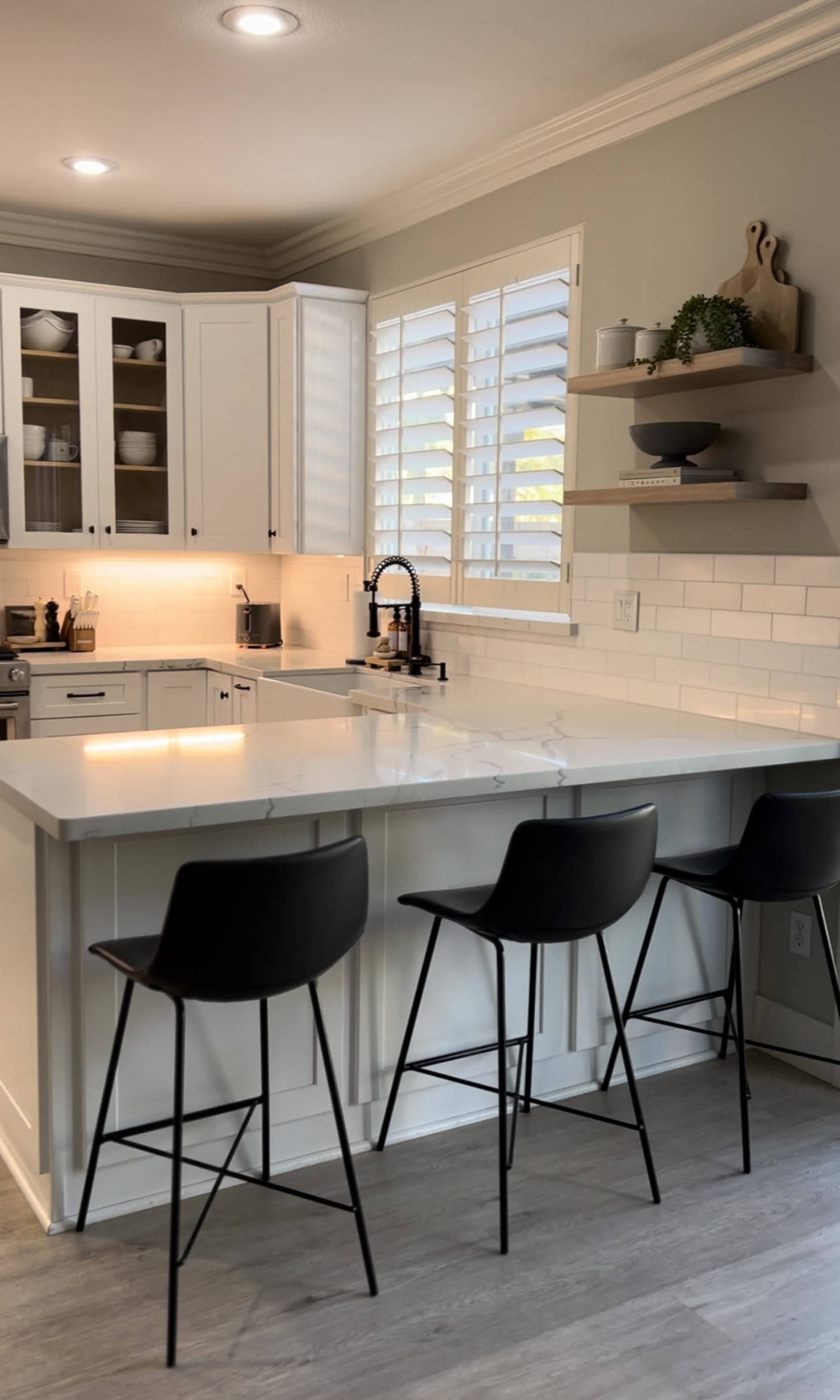 A modern kitchen features white cabinetry, a central island with marble countertops, and three black bar stools.