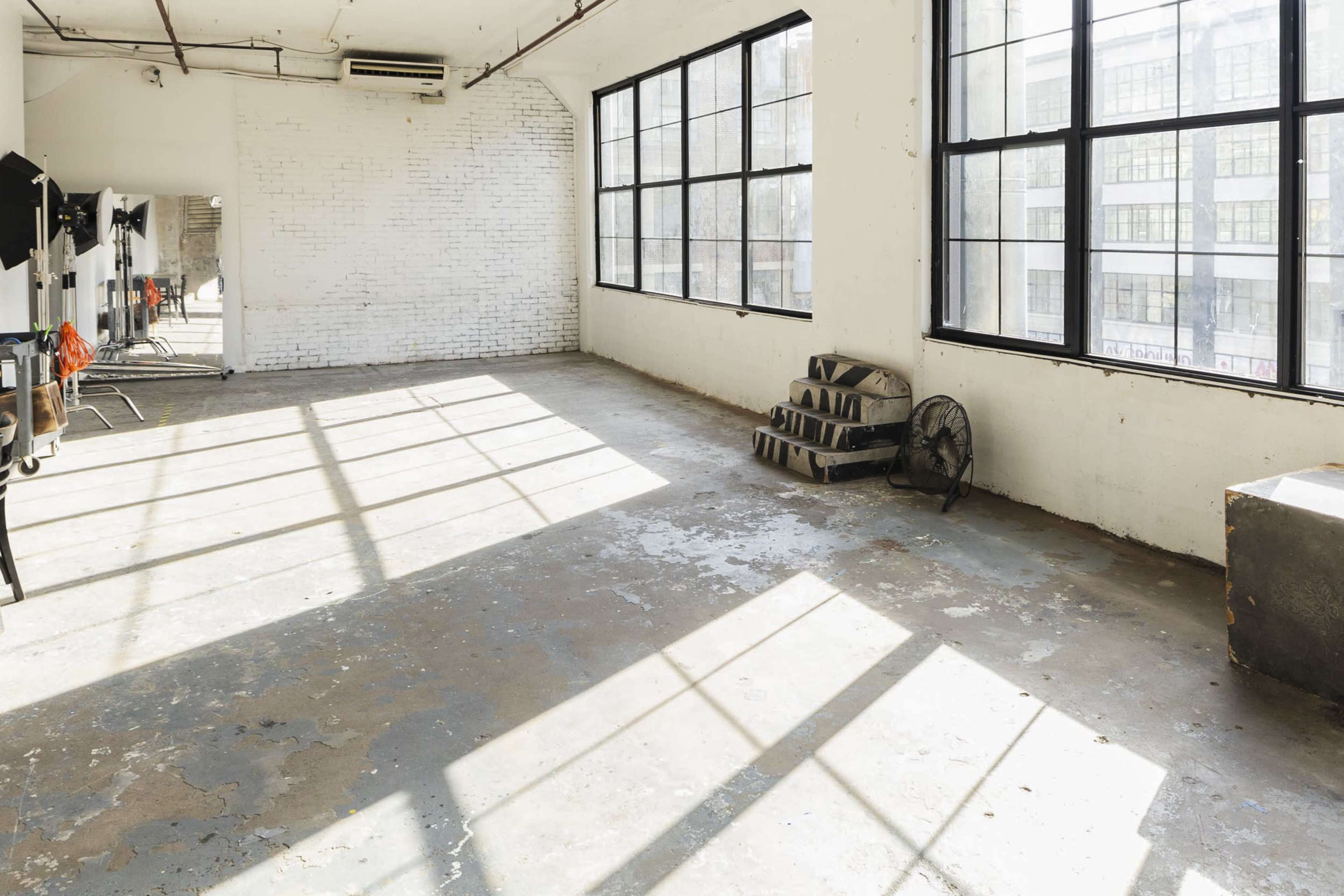 The image shows a spacious, sunlit room with large windows, a concrete floor, and a few scattered items including a fan and a stack of steps.