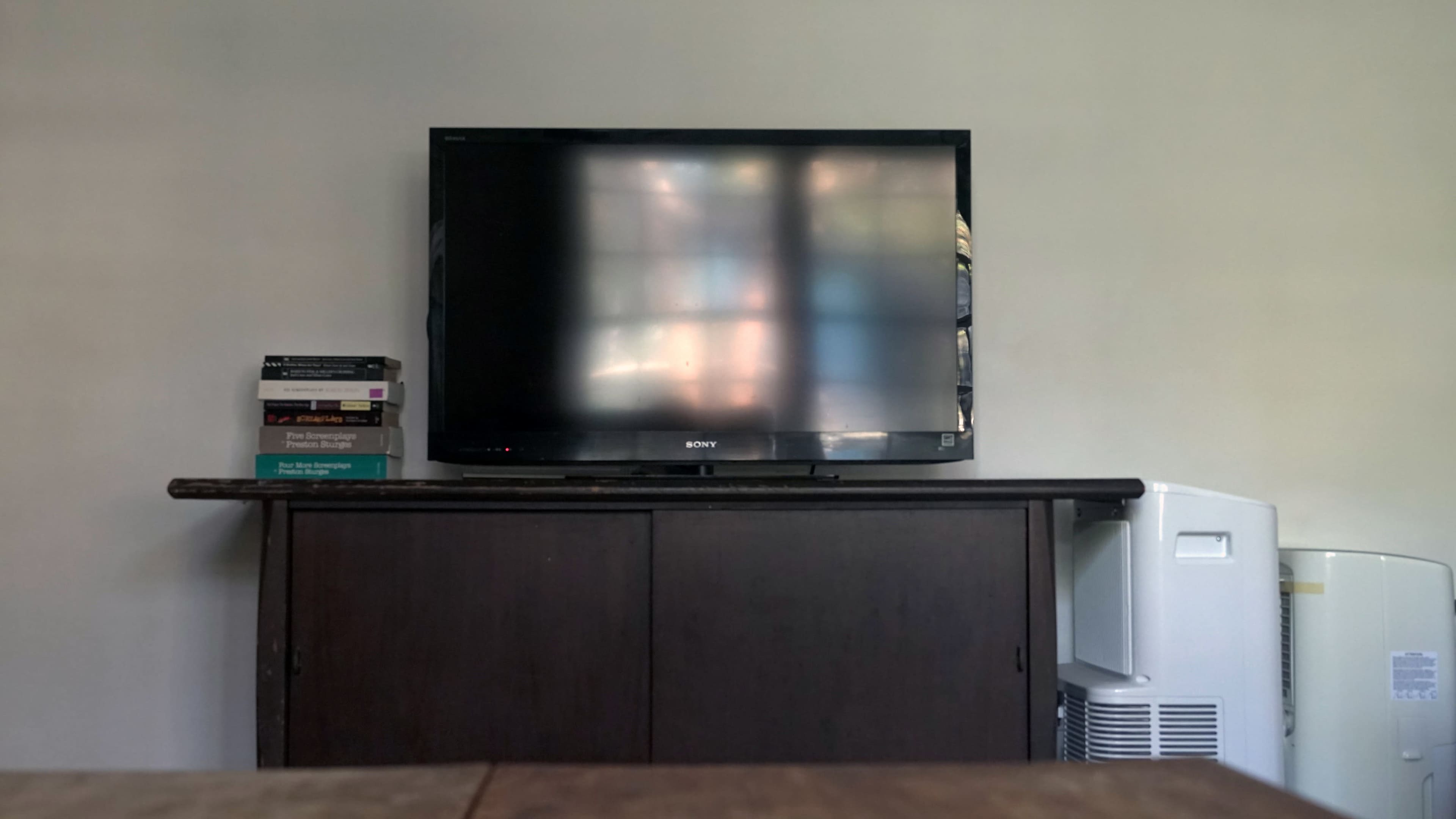 A television is mounted on a dark wooden cabinet next to a stack of books, with an air conditioner located to the right.