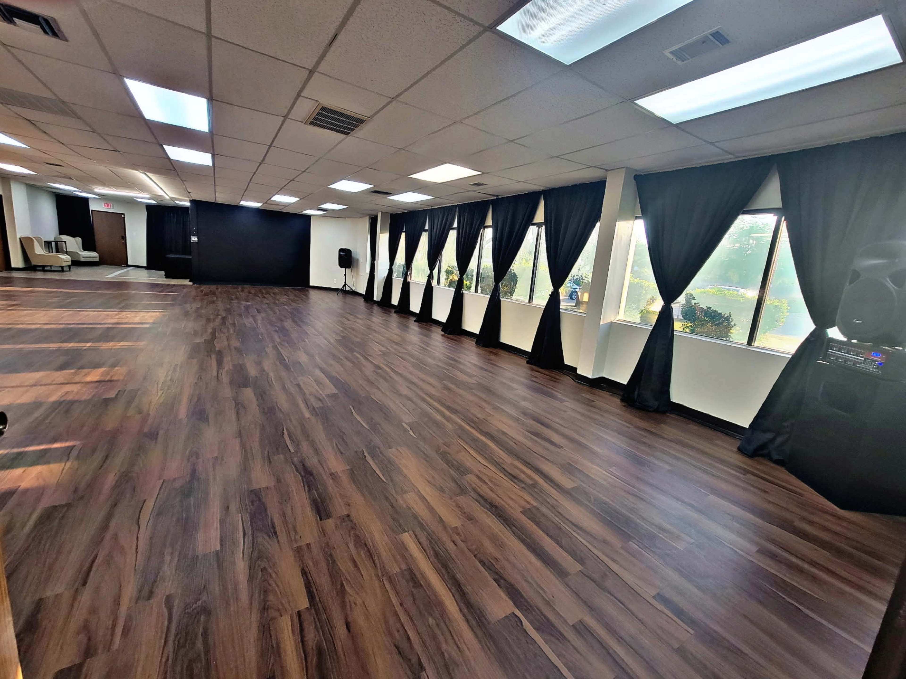 The image shows a spacious, well-lit studio with wooden flooring, featuring large windows covered with black curtains and a sound system in the corner.