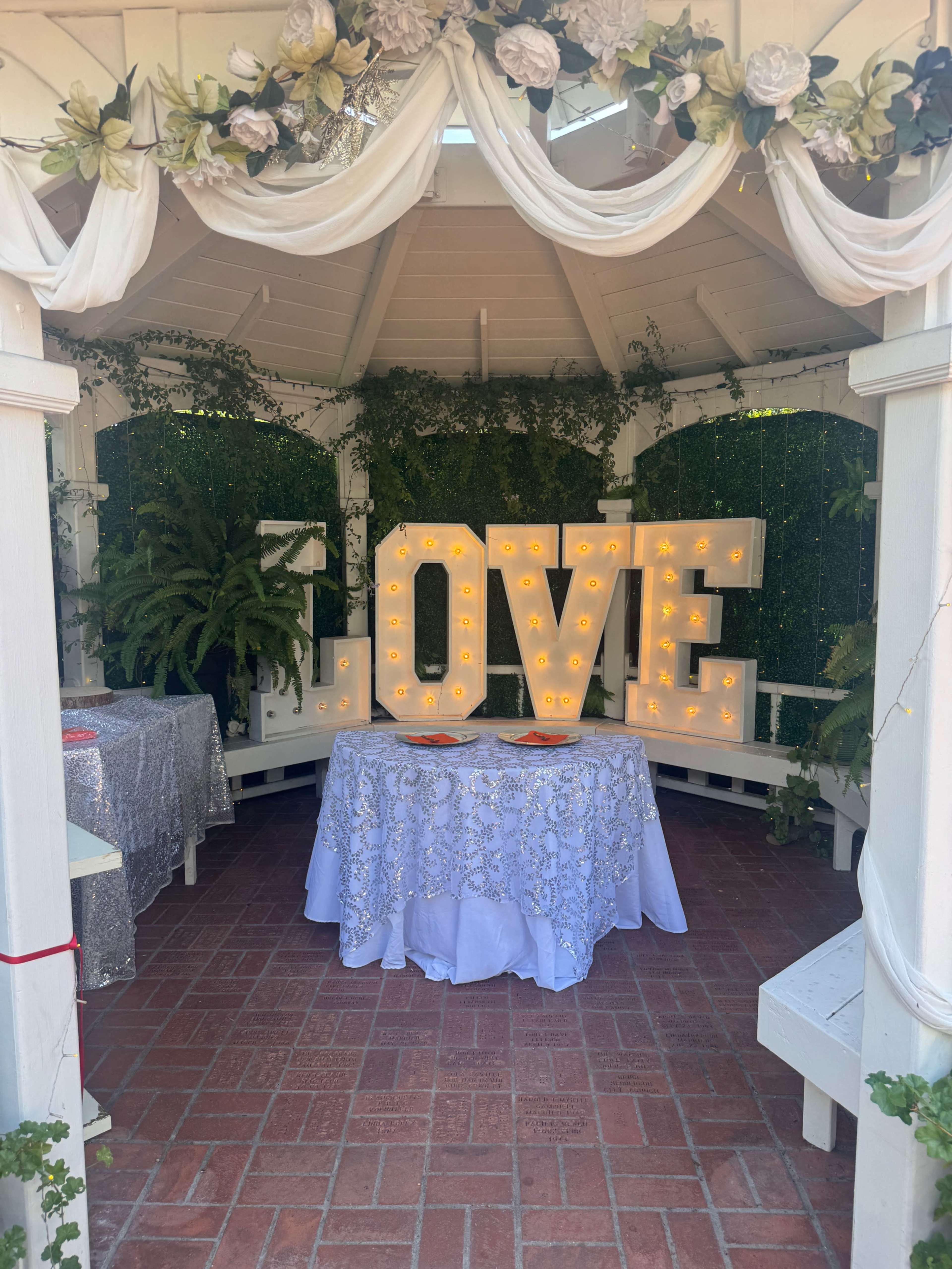 The image features a decorated gazebo with a large illuminated "LOVE" sign, surrounded by plants and a table covered in a lace tablecloth.