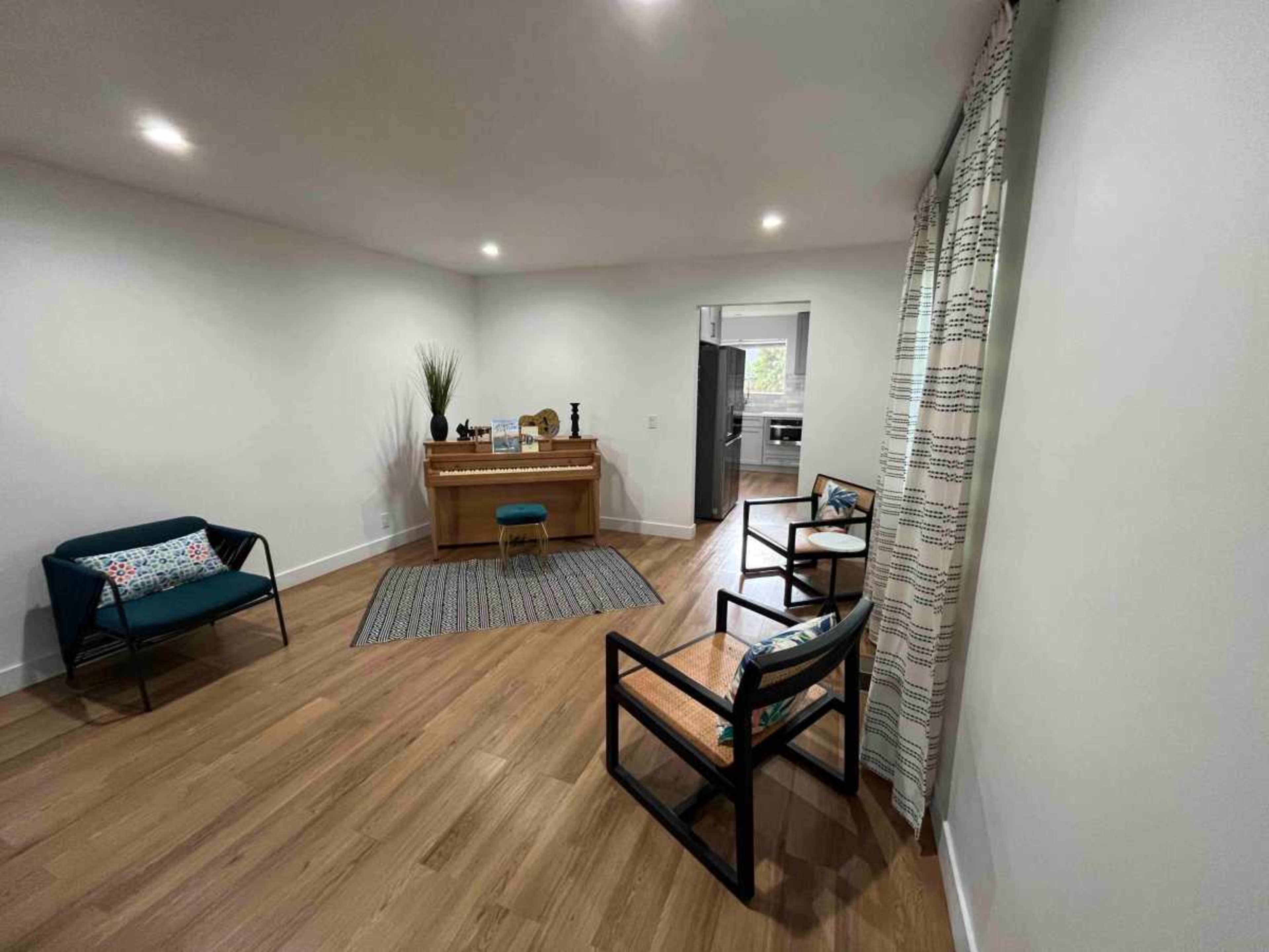 The image shows a sparsely furnished room with light-colored walls, wooden flooring, and a small table, accompanied by three chairs and a rug.