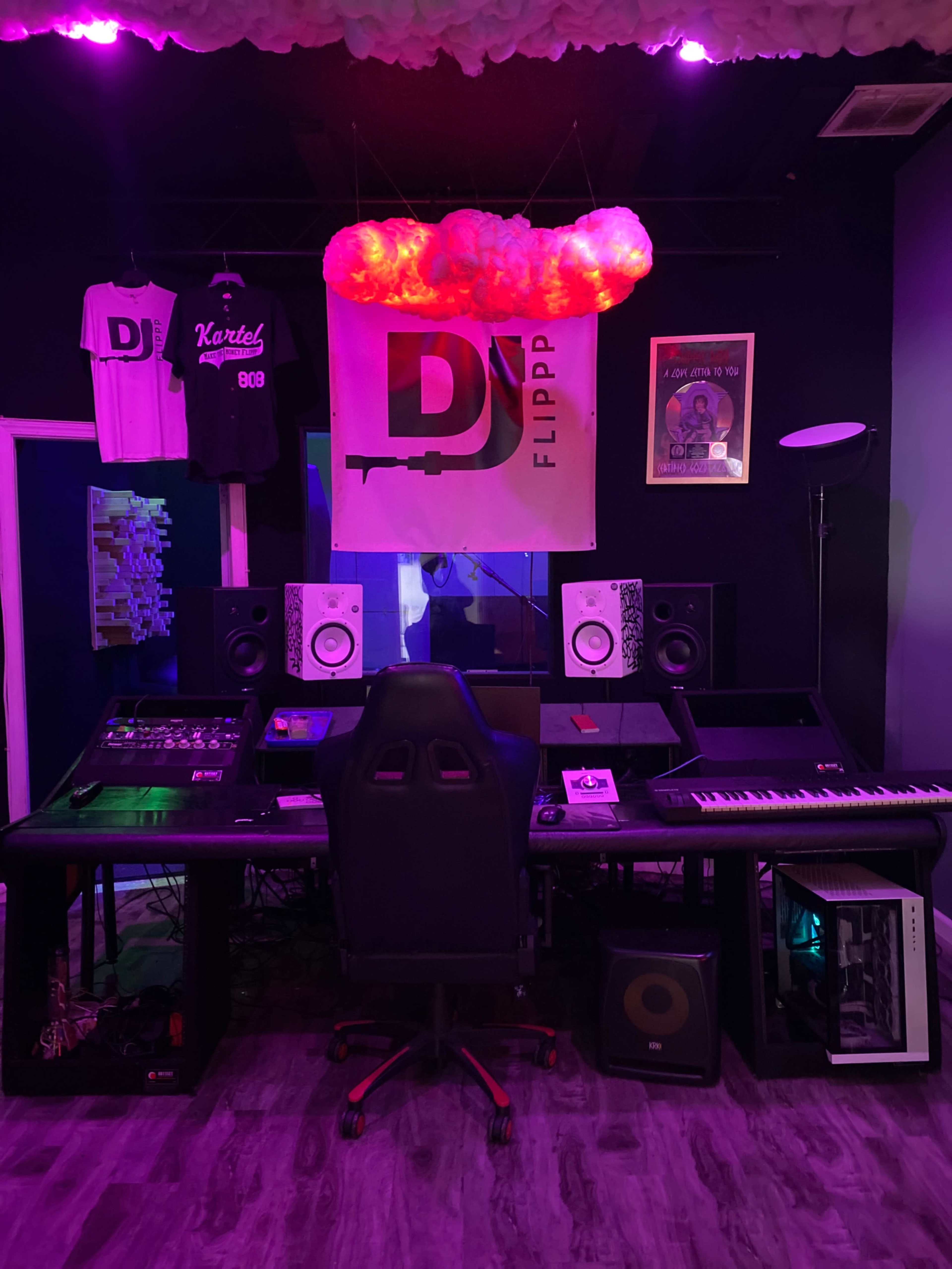 A music studio with a desk equipped with audio equipment, speakers, a keyboard, and DJ-related decor on the walls.