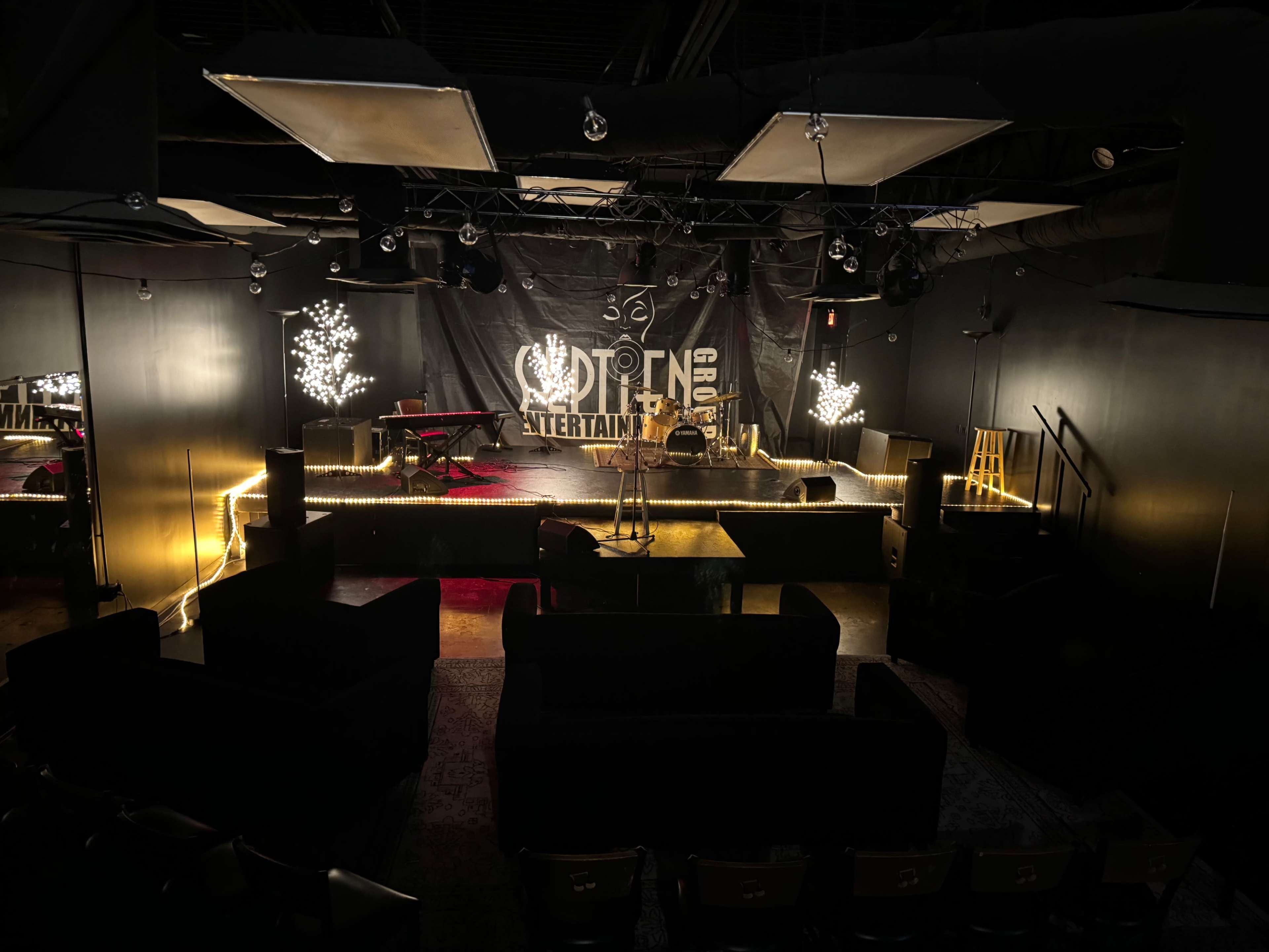 A stage area in a dark venue, featuring a microphone stand, a drum set, and seating arrangements with couches and chairs.