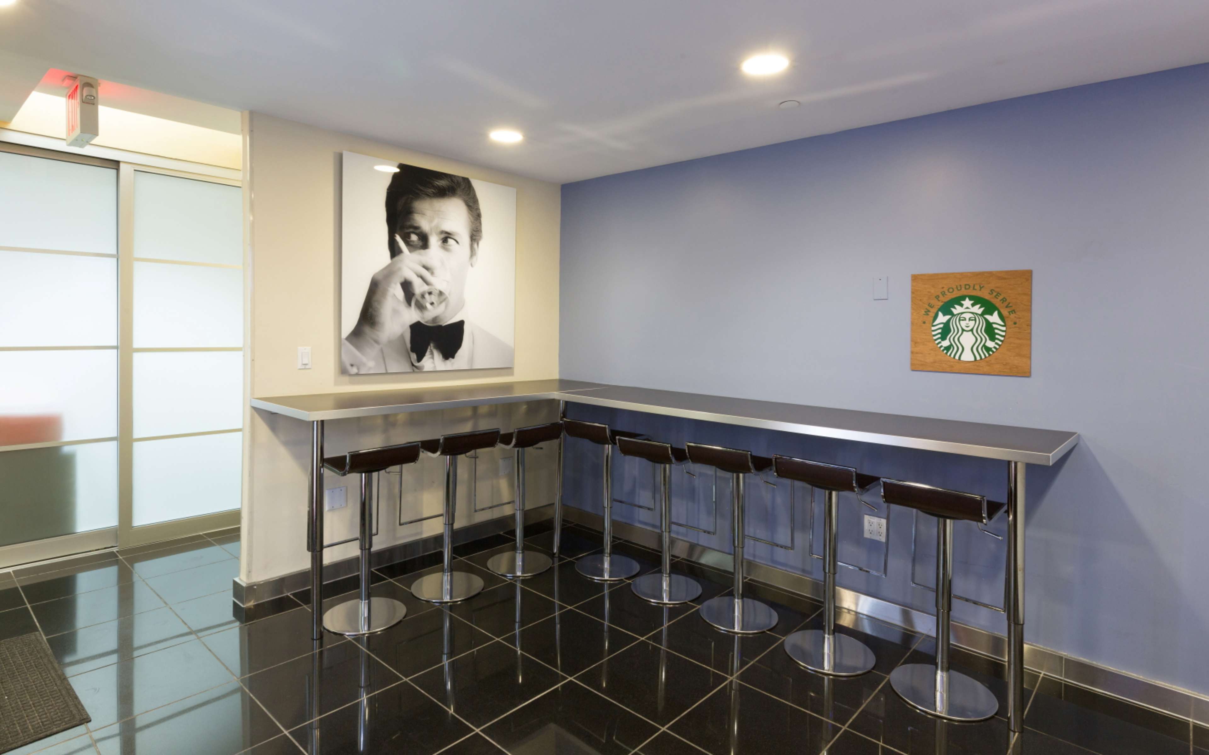 A modern lounge area features a metal countertop with high stools and a large black-and-white portrait of a man drinking coffee on the wall.