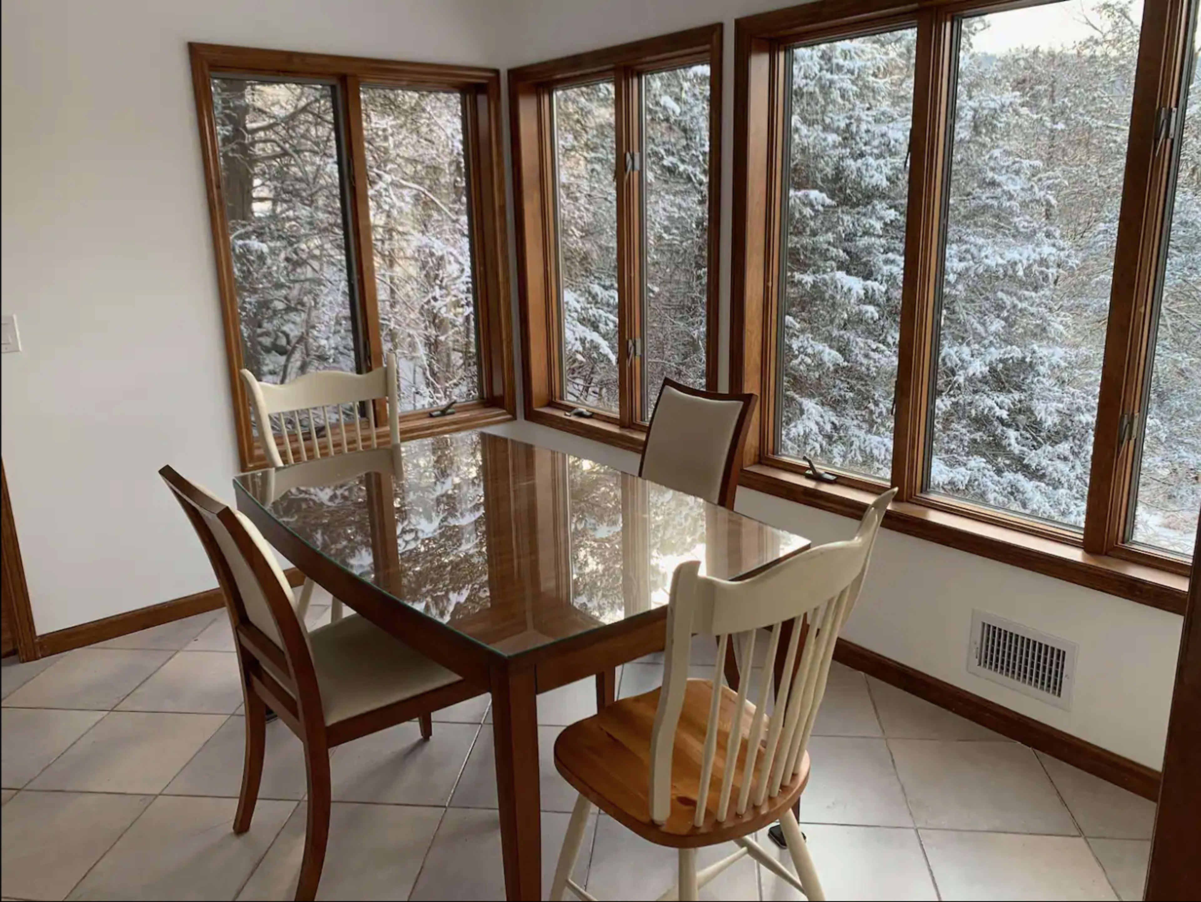 A sunlit dining area features a glass table surrounded by four chairs, with large windows providing a view of snowy trees outside.
