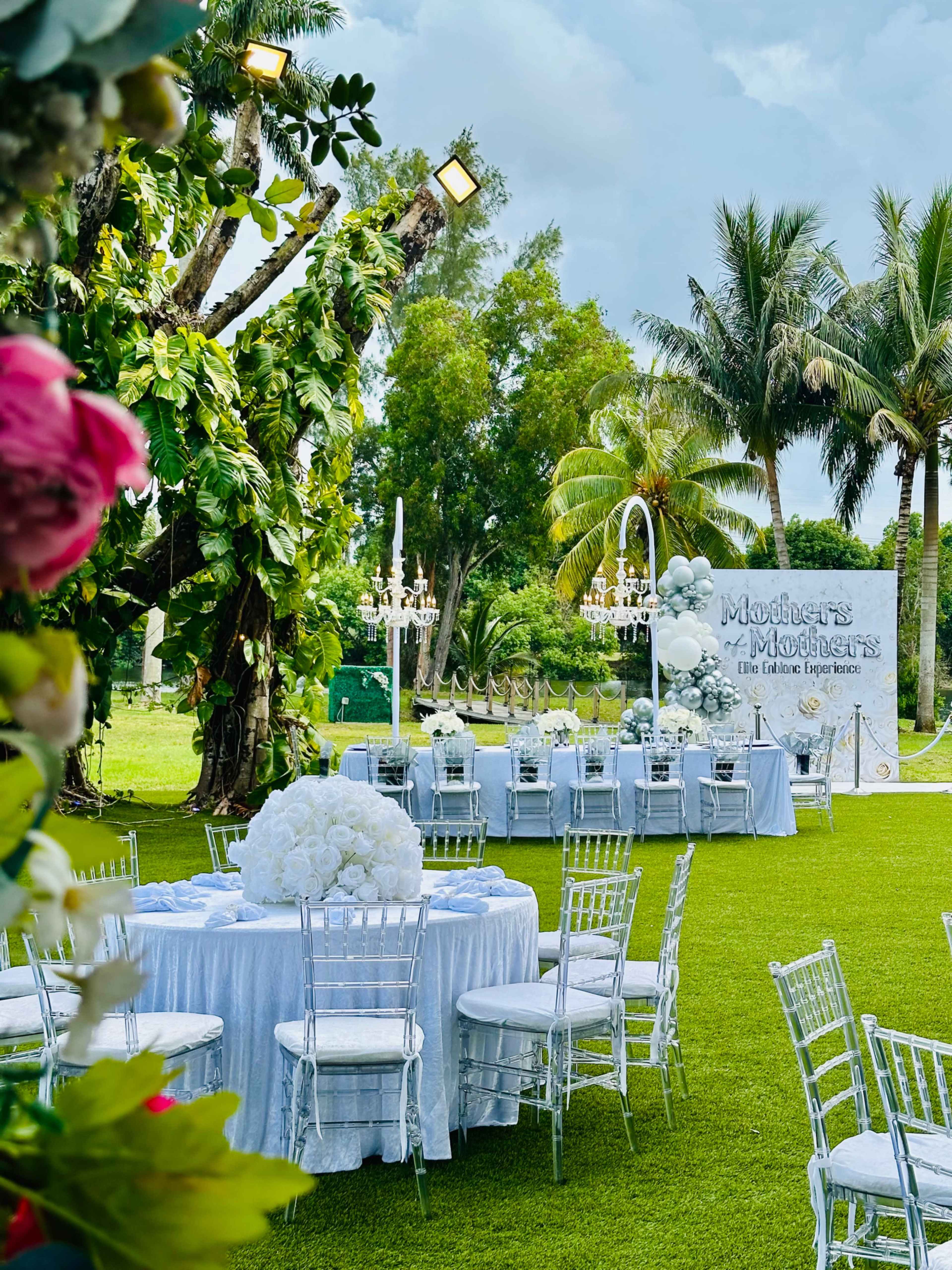 A beautifully arranged outdoor event space features round tables with white tablecloths and large floral centerpieces, surrounded by greenery and palm trees.