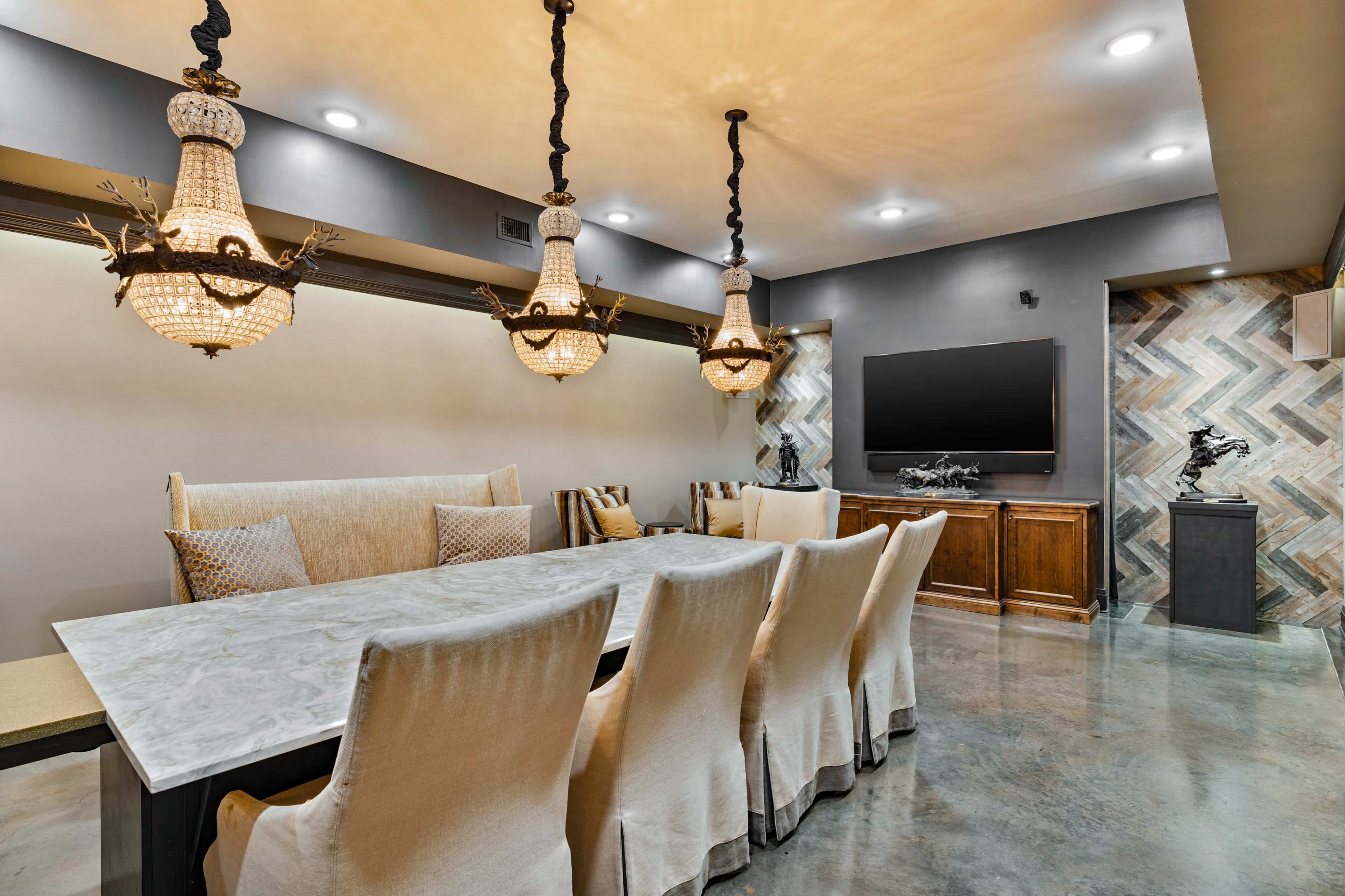 The image shows a modern dining room with a large table surrounded by upholstered chairs, illuminated by ornate chandeliers, and featuring a wall-mounted TV.