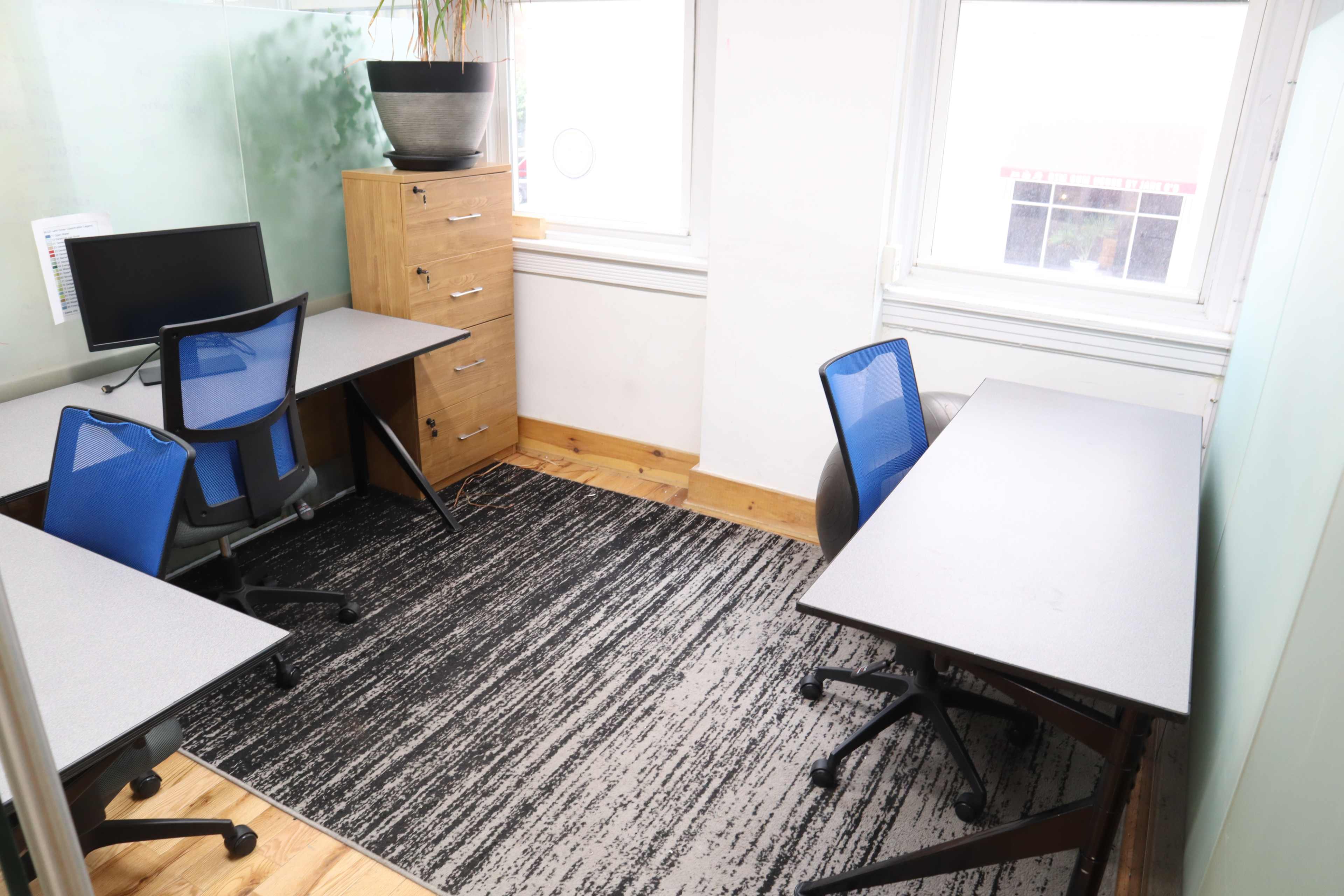 A small office space with two desks, chairs, and a storage unit, illuminated by natural light from windows.