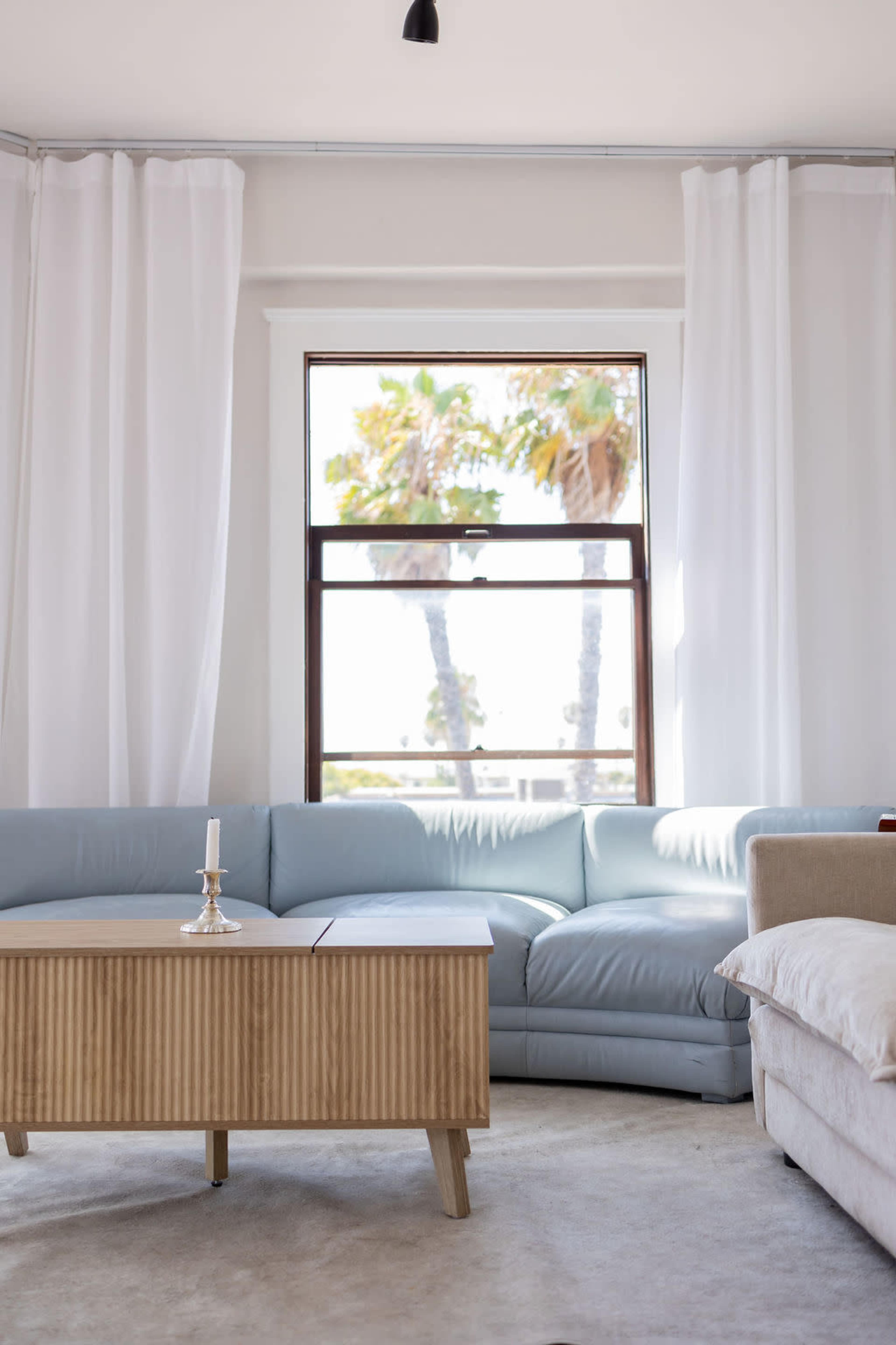 The image shows a light-colored living room with a blue sectional sofa and a wooden coffee table, featuring large windows with sheer curtains and palm trees visible outside.