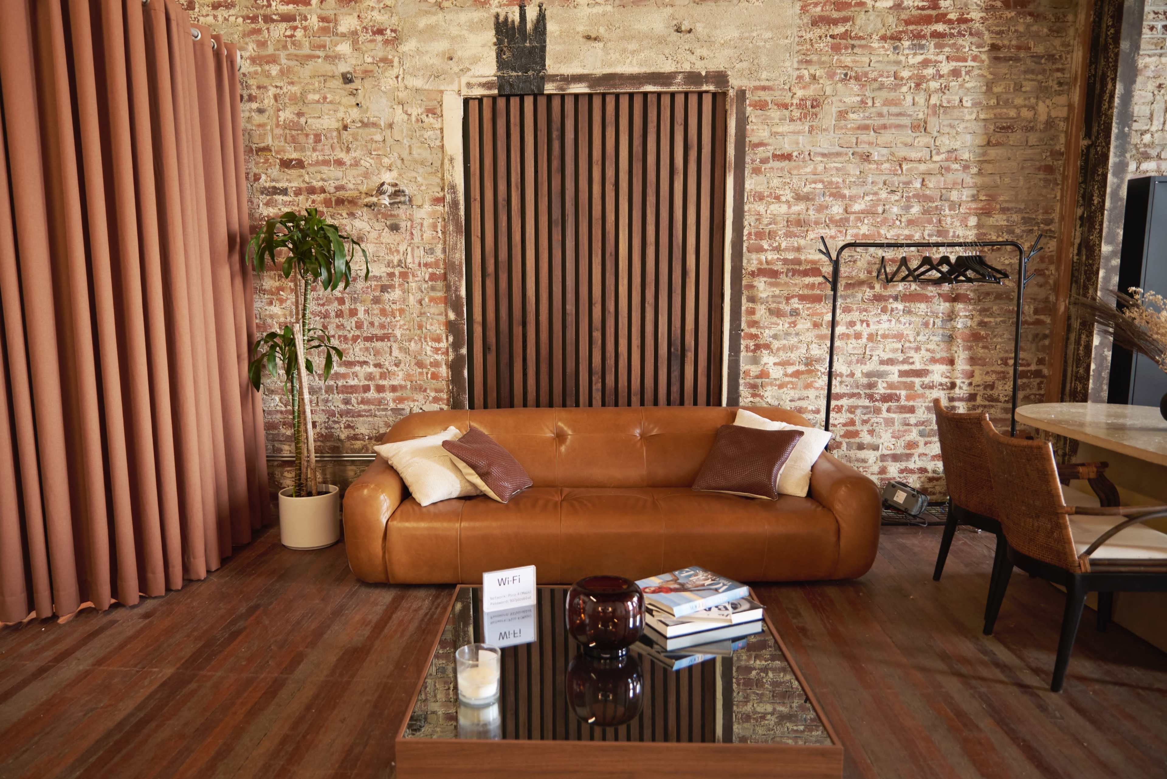 The image shows a modern living space with a brown leather sofa, a wooden coffee table with books and a decorative item, and a backdrop of exposed brick walls and wooden slatted doors.