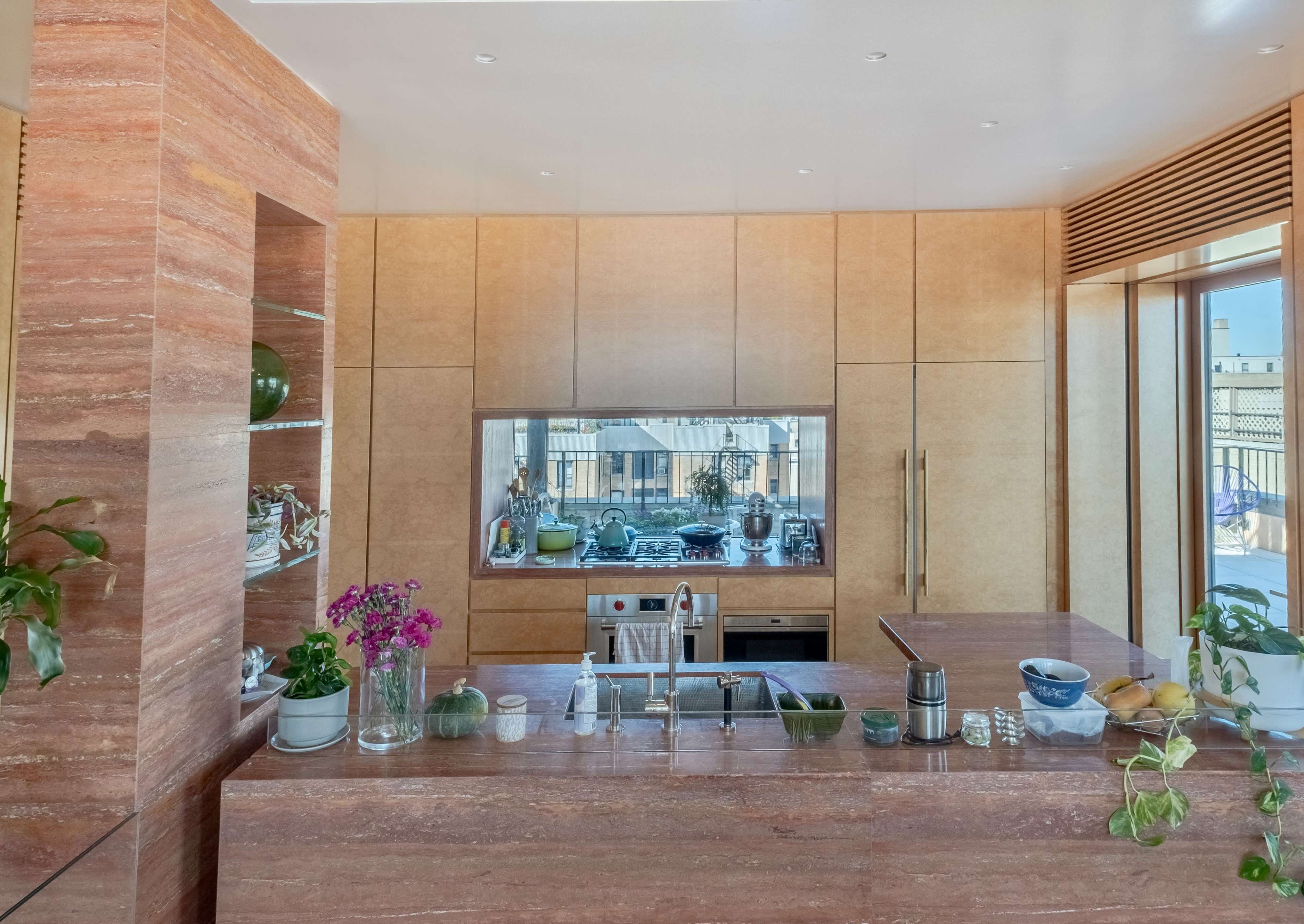 A modern kitchen features a stone island, a sleek counter, and a window overlooking a balcony with various plants.