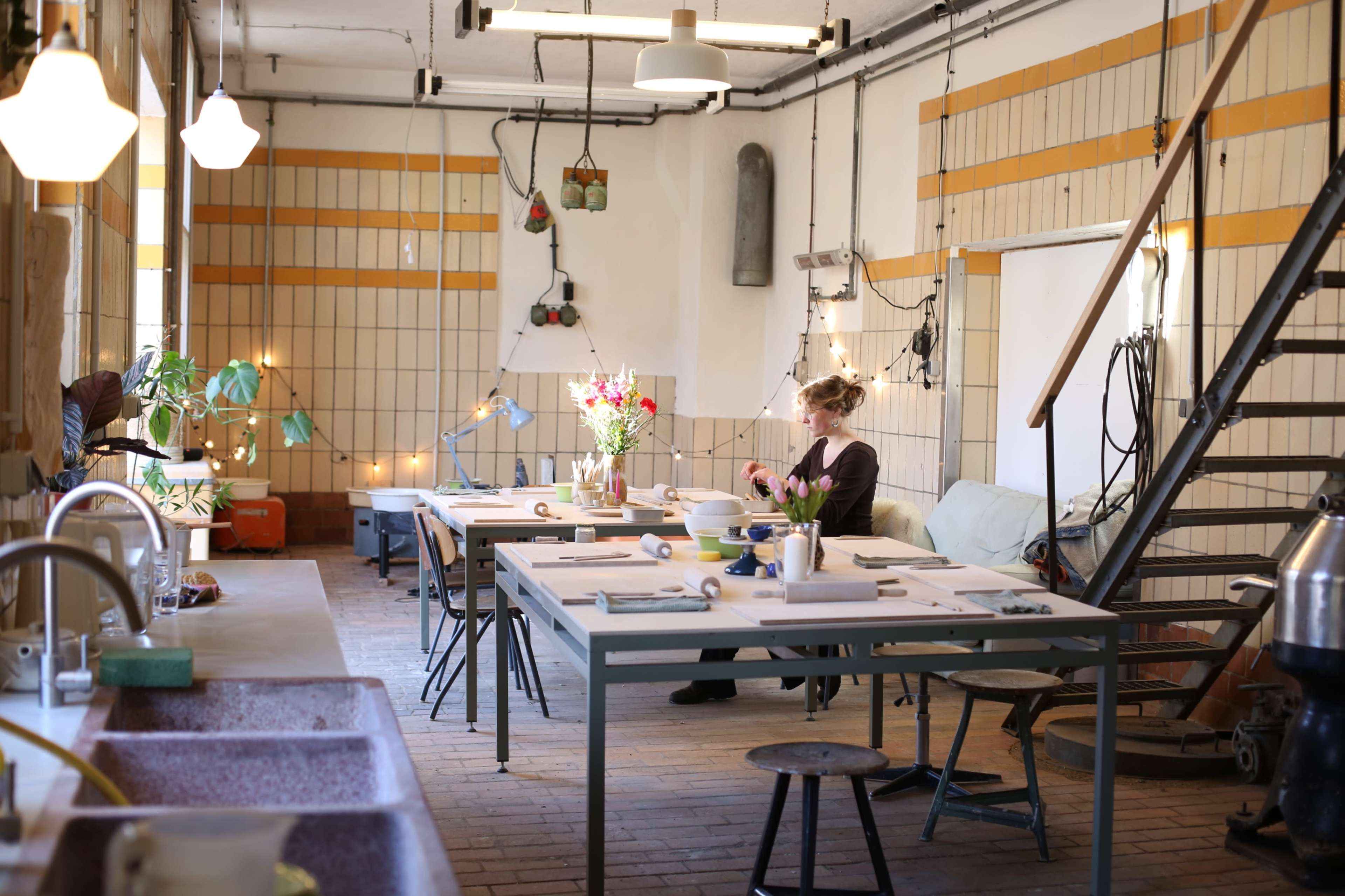 A person sits at a table in a spacious, well-lit studio filled with plants and art supplies, while another table is arranged for creative work.