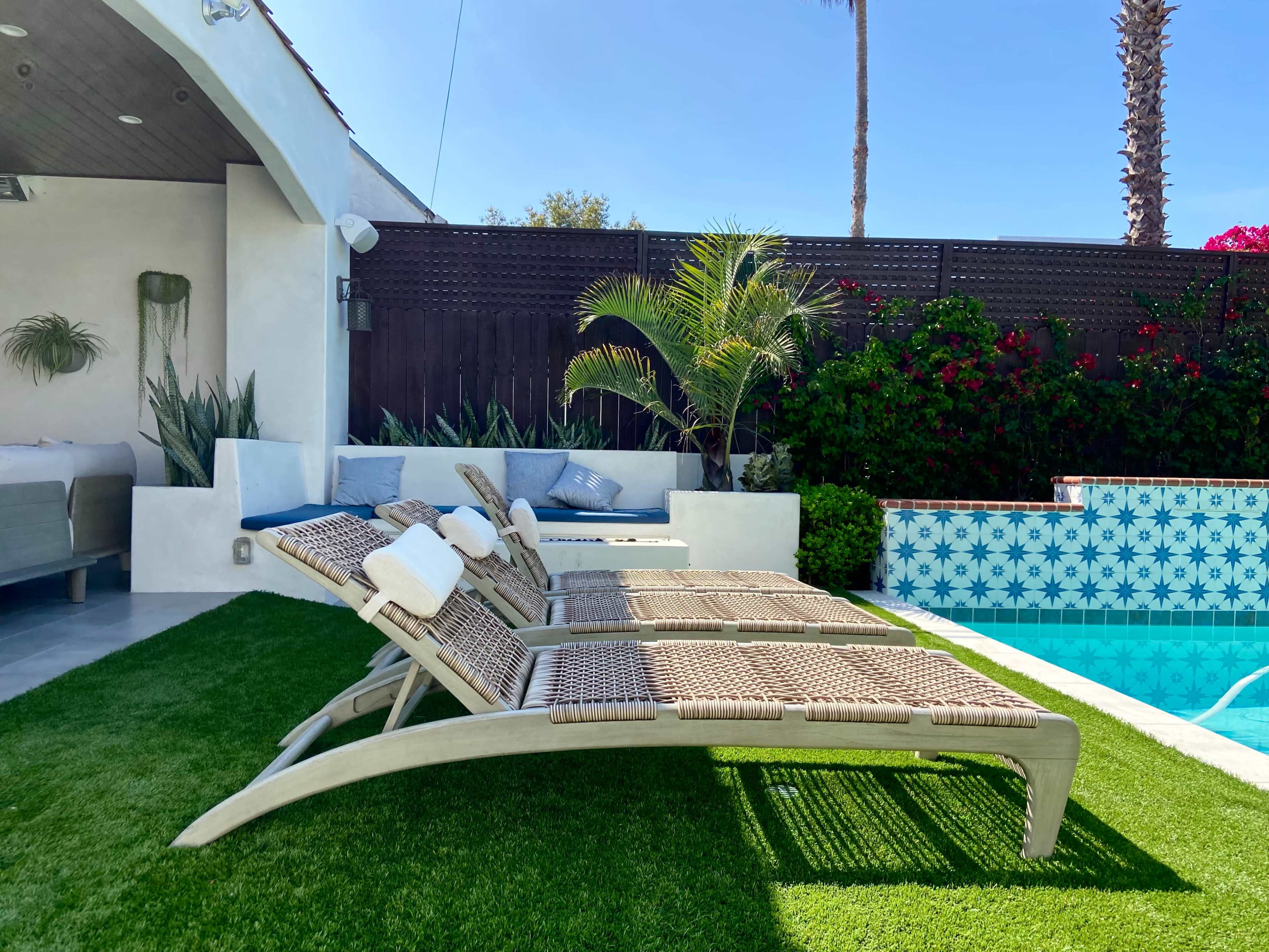 Two loungers are positioned beside a swimming pool surrounded by vibrant greenery and patterned tiles.
