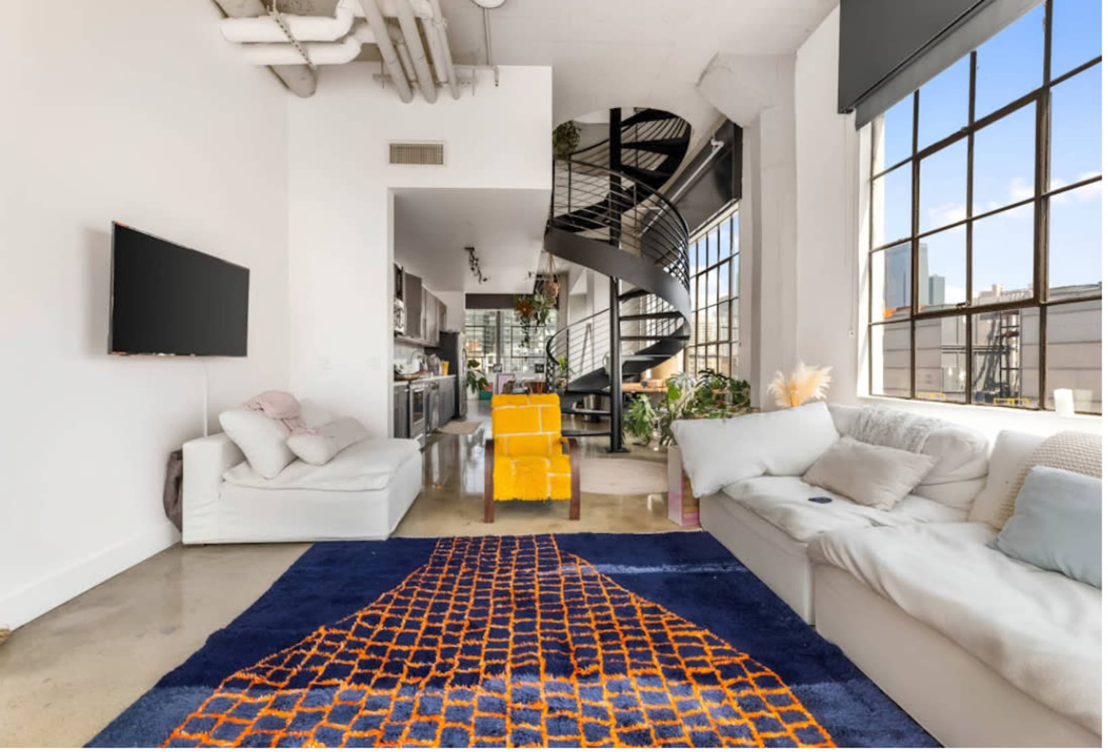 The image shows a modern, open-concept living space featuring a light-colored sofa, a vibrant rug, a yellow accent chair, and a spiral staircase leading to a loft area.