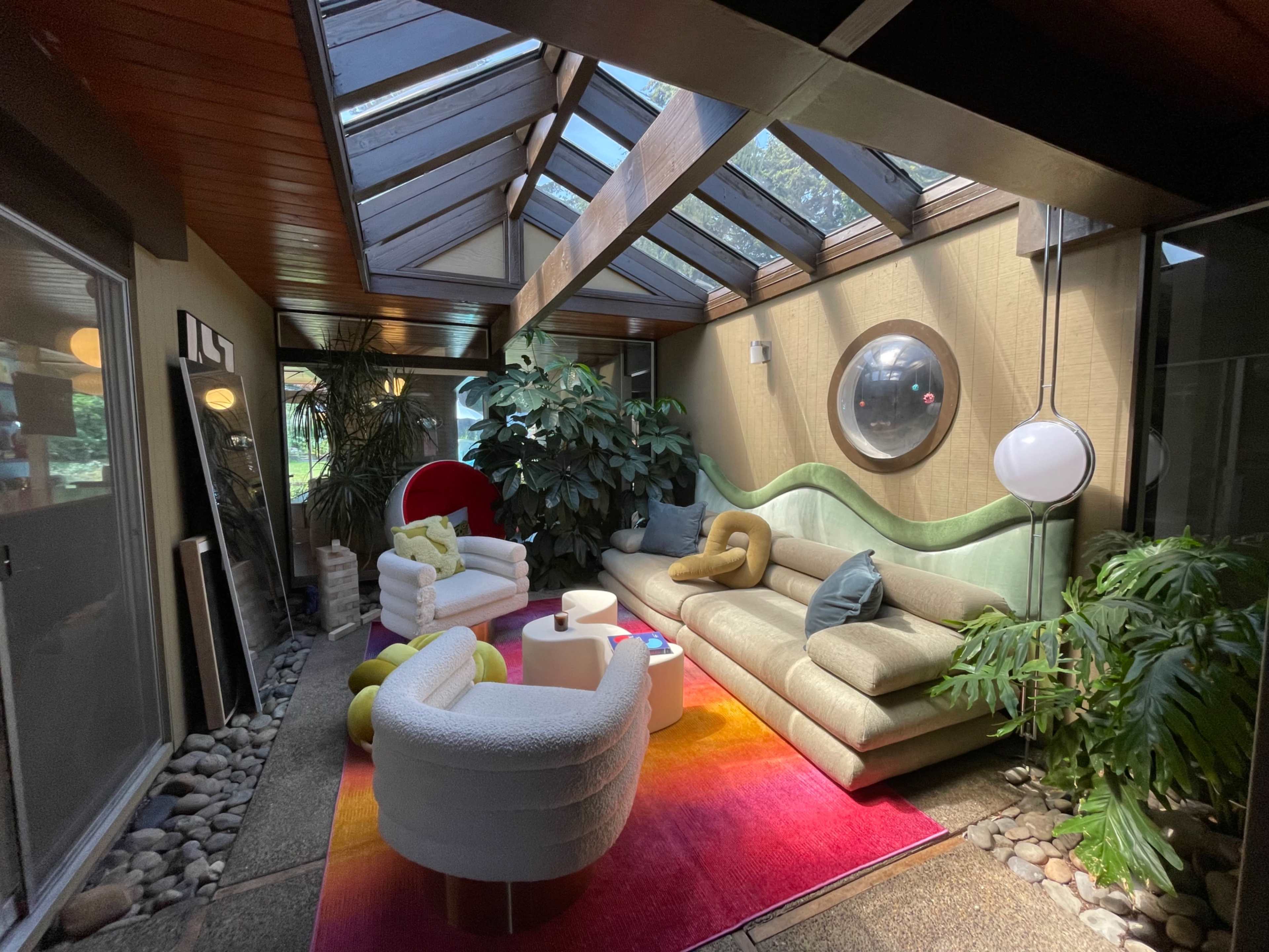 The image shows a modern sunlit living space with large skylights, a colorful rug, and various seating arrangements surrounded by indoor plants.