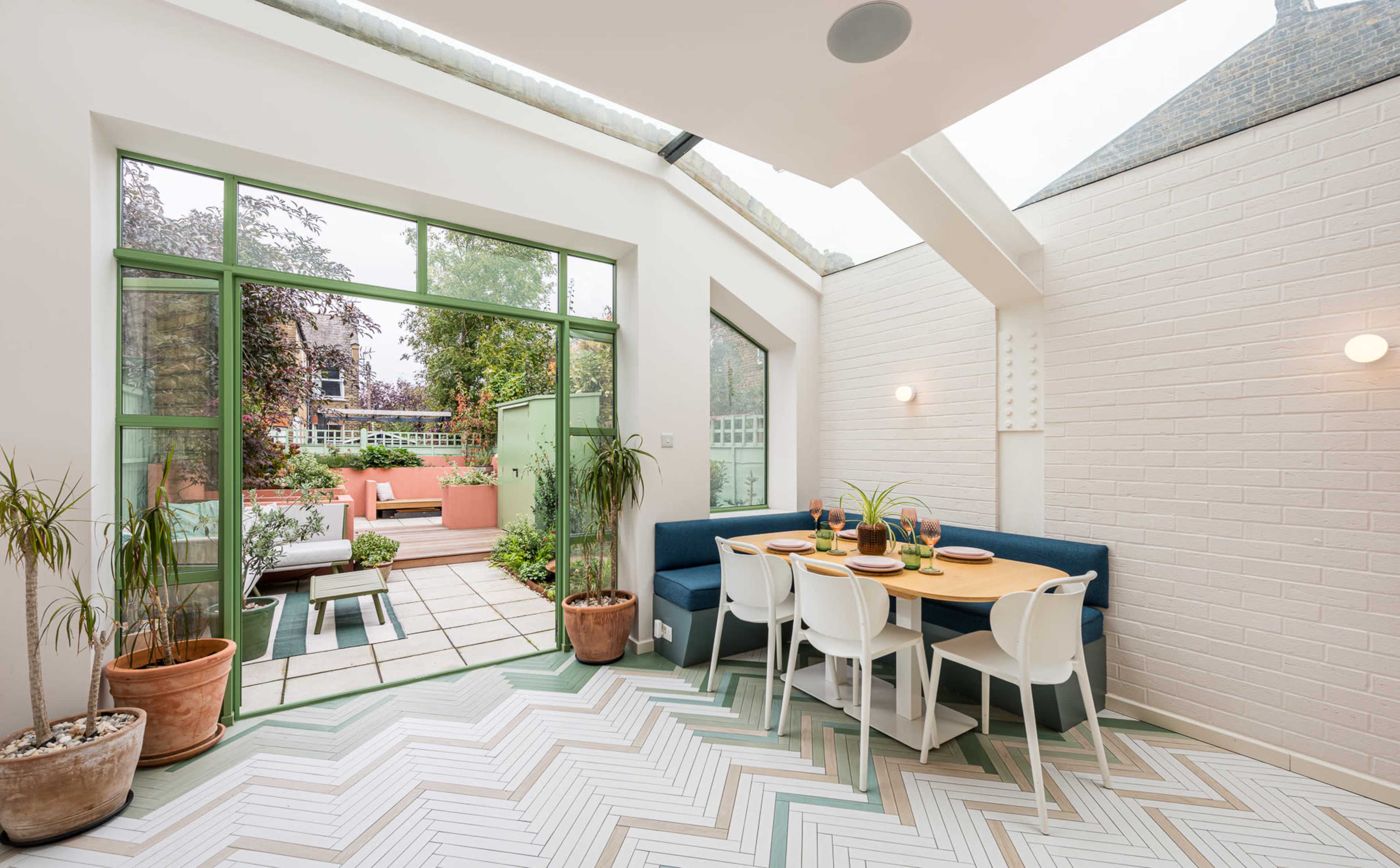 The image shows a bright, modern dining area with a blue bench seating, a table set for a meal, and large windows opening to a light-filled garden.