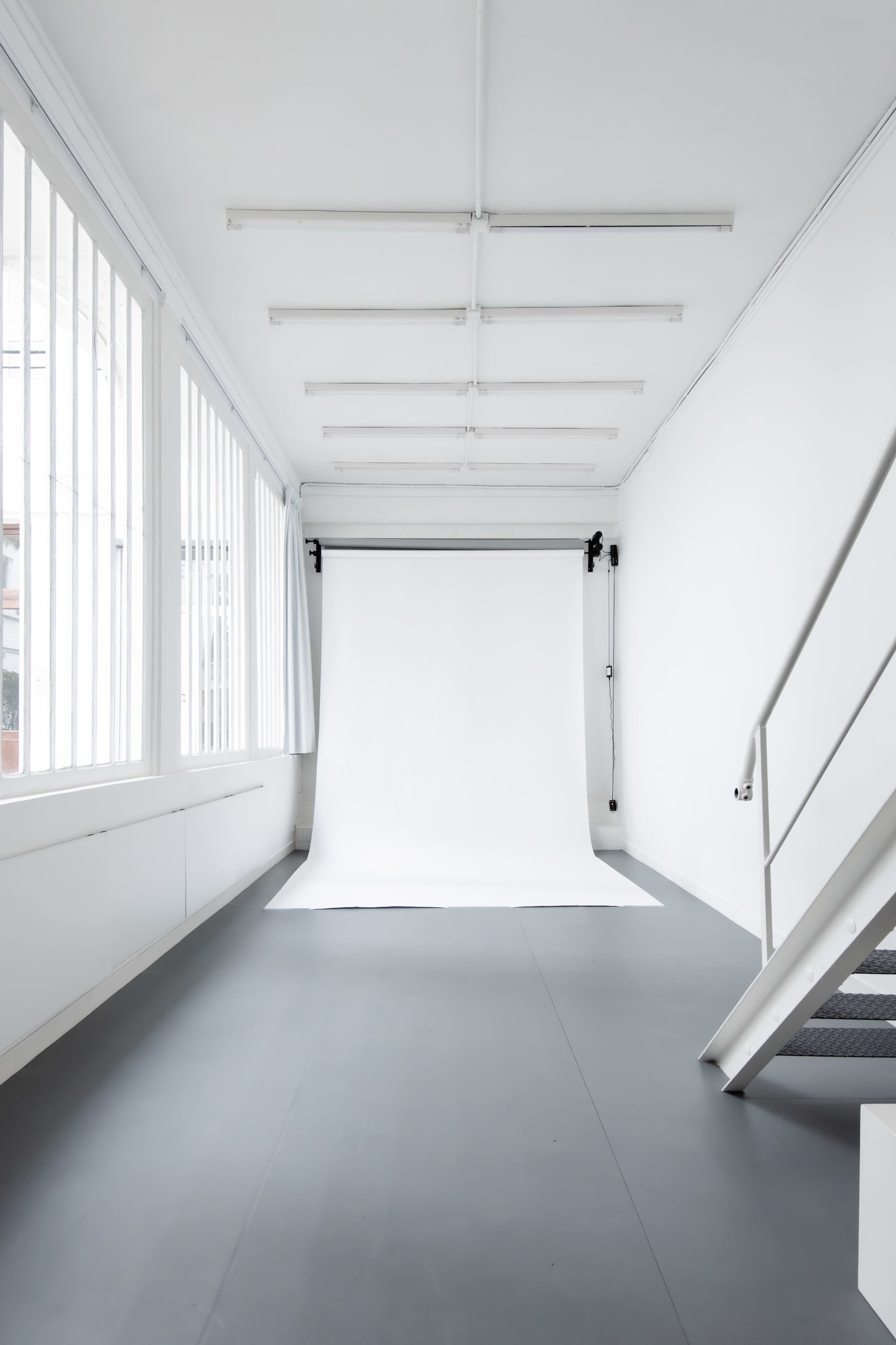 The image shows a spacious white studio with a gray floor and a white backdrop, illuminated by natural light through large windows.