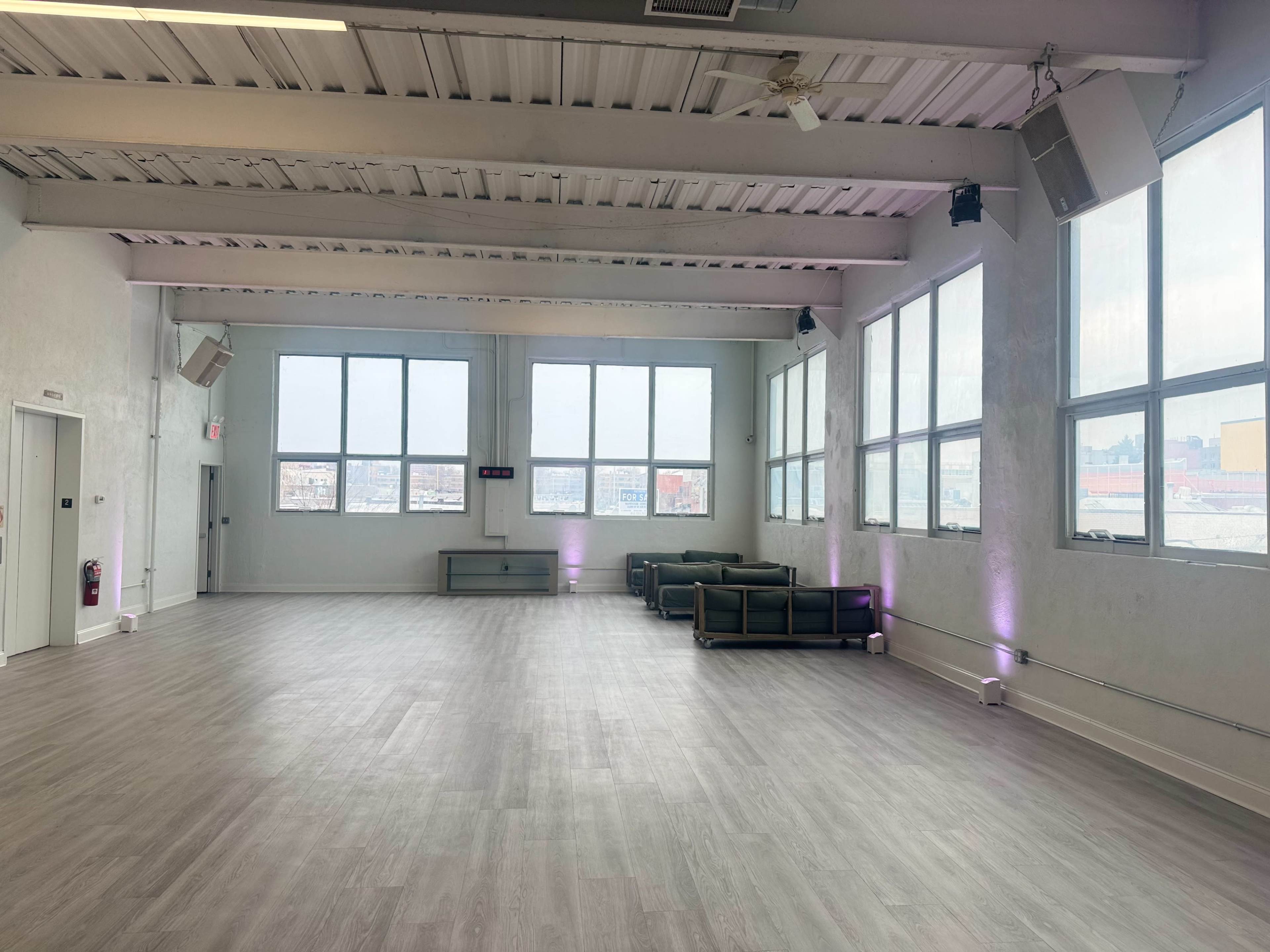 The image shows a spacious, modern interior with large windows, gray flooring, and black sofas arranged along one side of the room.