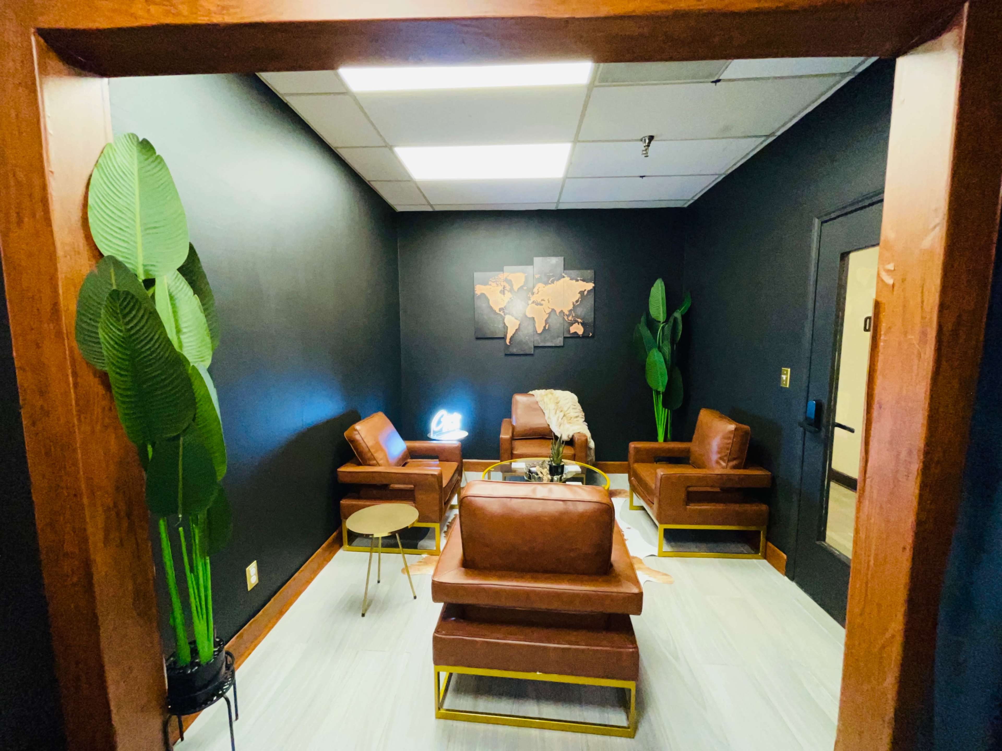 A modern lounge area with brown leather chairs, a small circular table, and potted plants, all set against a black wall featuring a world map.