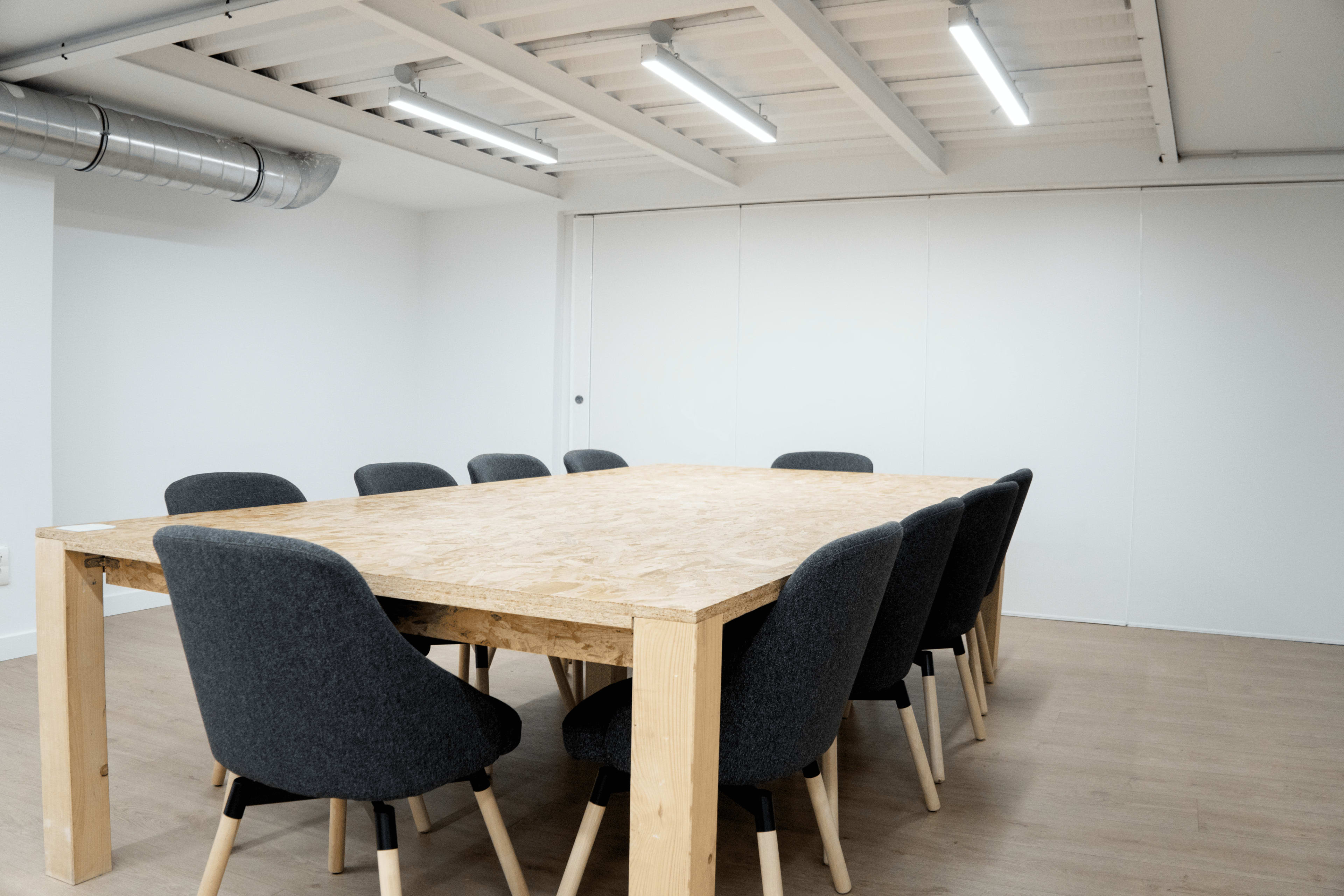 A large, rectangular wooden table is surrounded by eight upholstered chairs in a brightly lit meeting room.