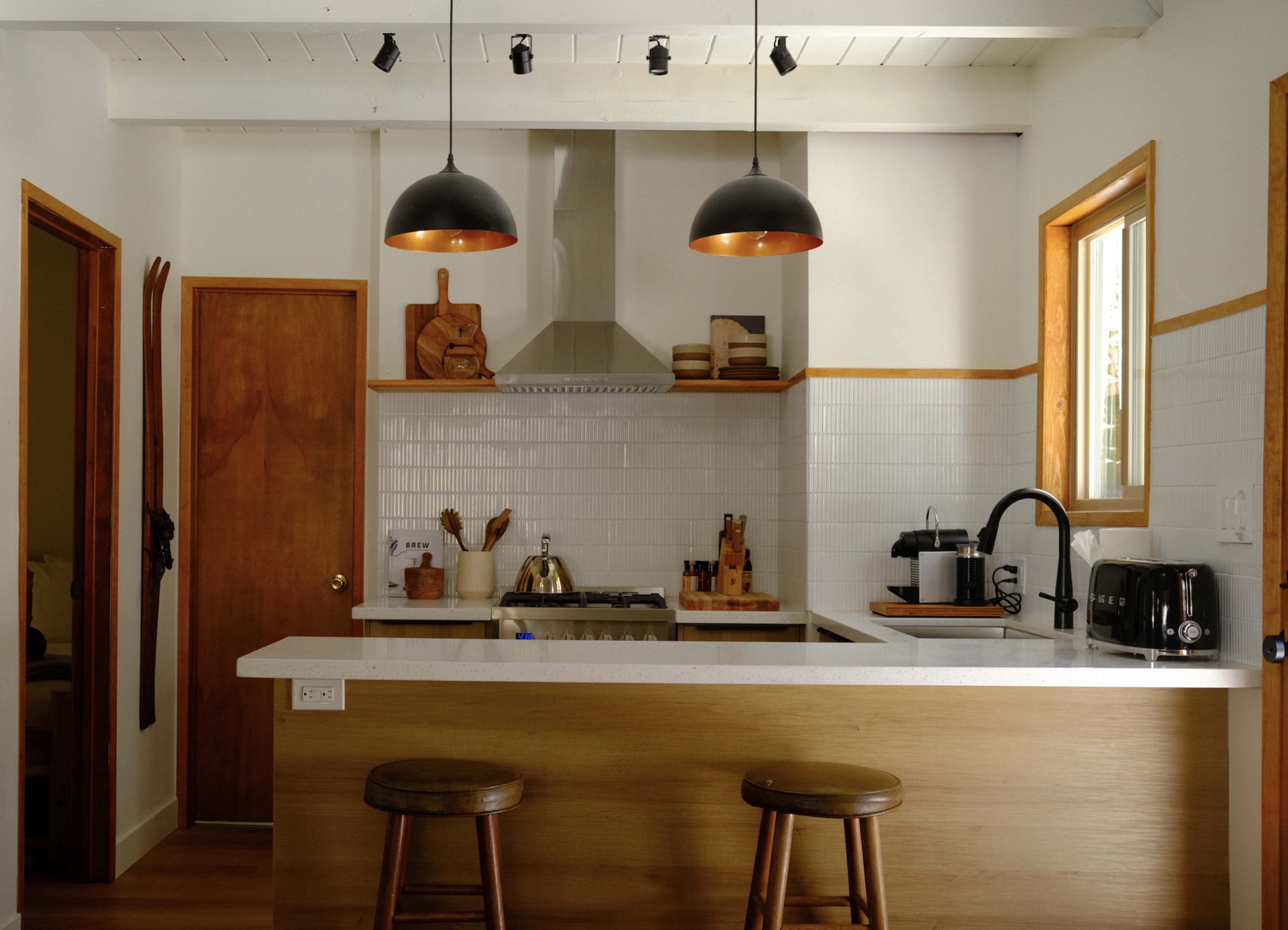The image shows a modern kitchen with a wooden bar, two stools, stainless steel appliances, and black pendant lights.