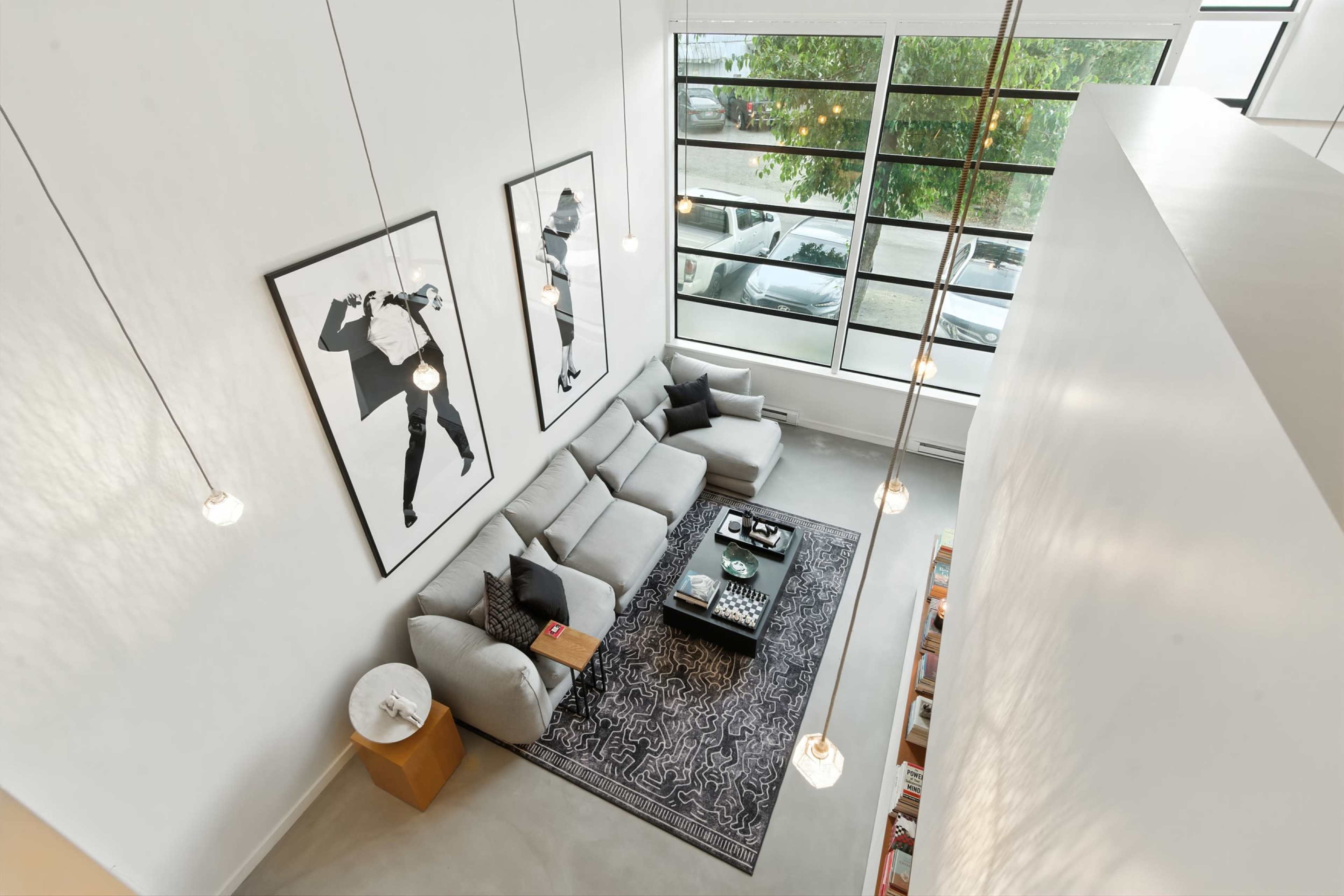 A modern, open-concept living room features a large gray sectional sofa, two framed black-and-white artworks, and a black coffee table on a patterned rug, with large windows letting in natural light.