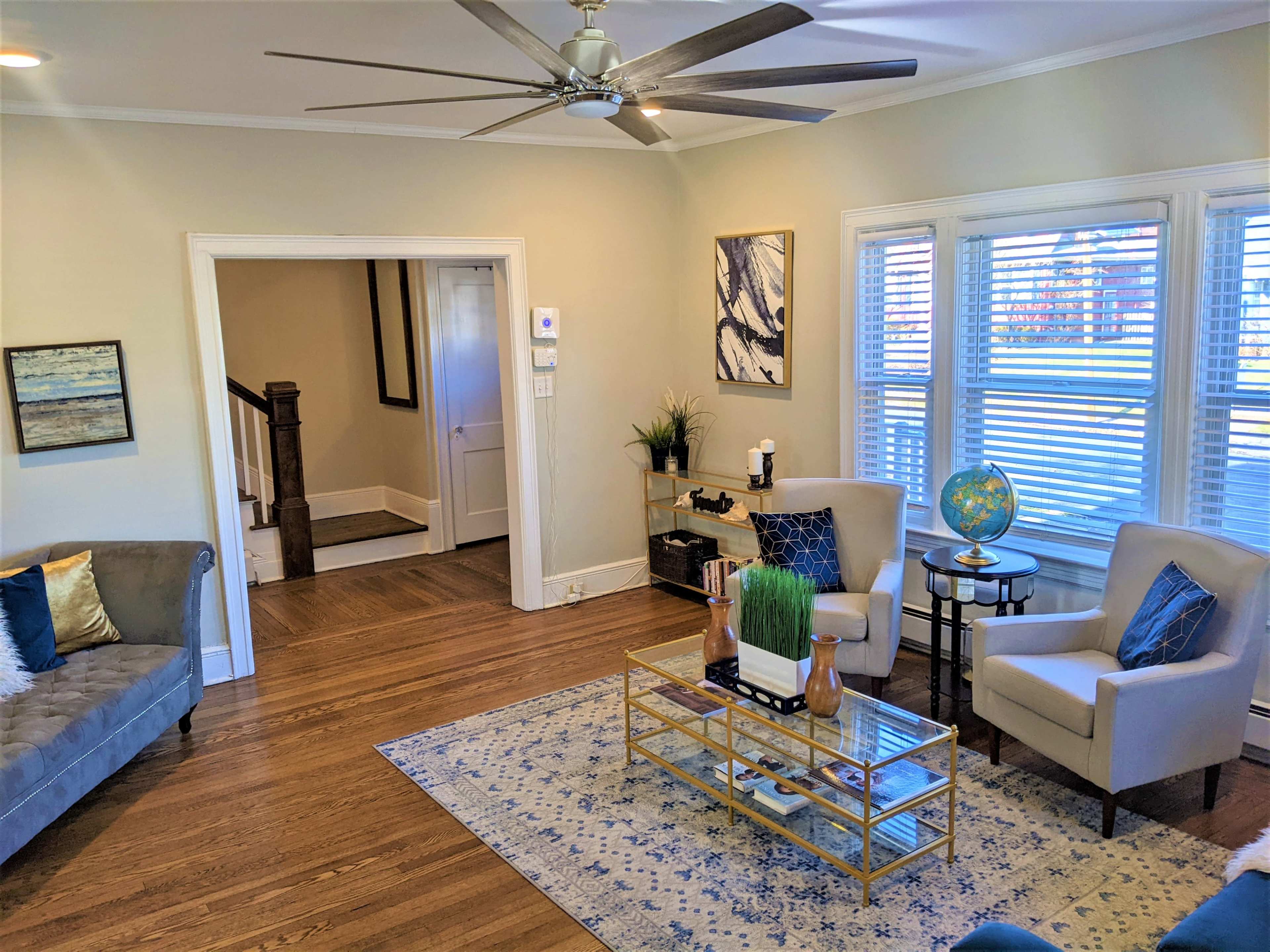 A cozy living room with wooden floors, a gray couch, two cream-colored chairs, and a coffee table on a patterned rug, complemented by natural light coming through the window.