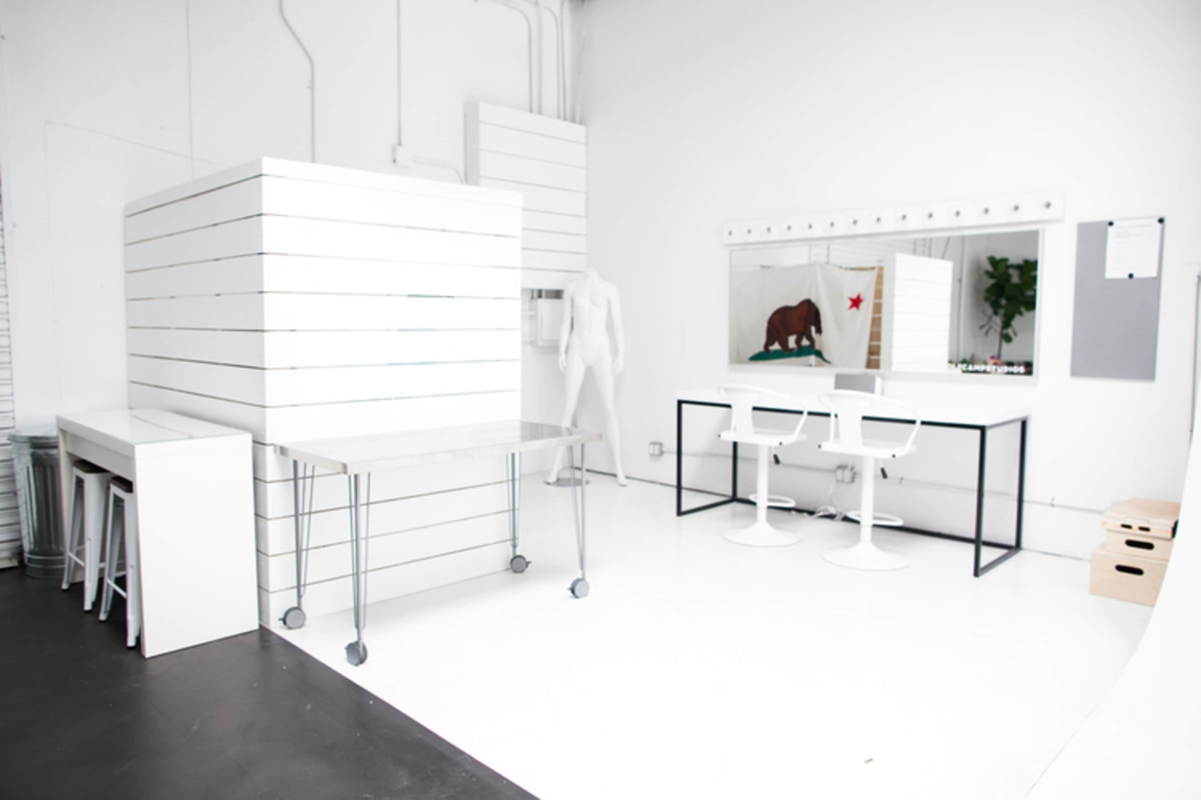A modern, minimalist studio with white walls, a large mirror, a table on wheels, and a display area with slatted surfaces.