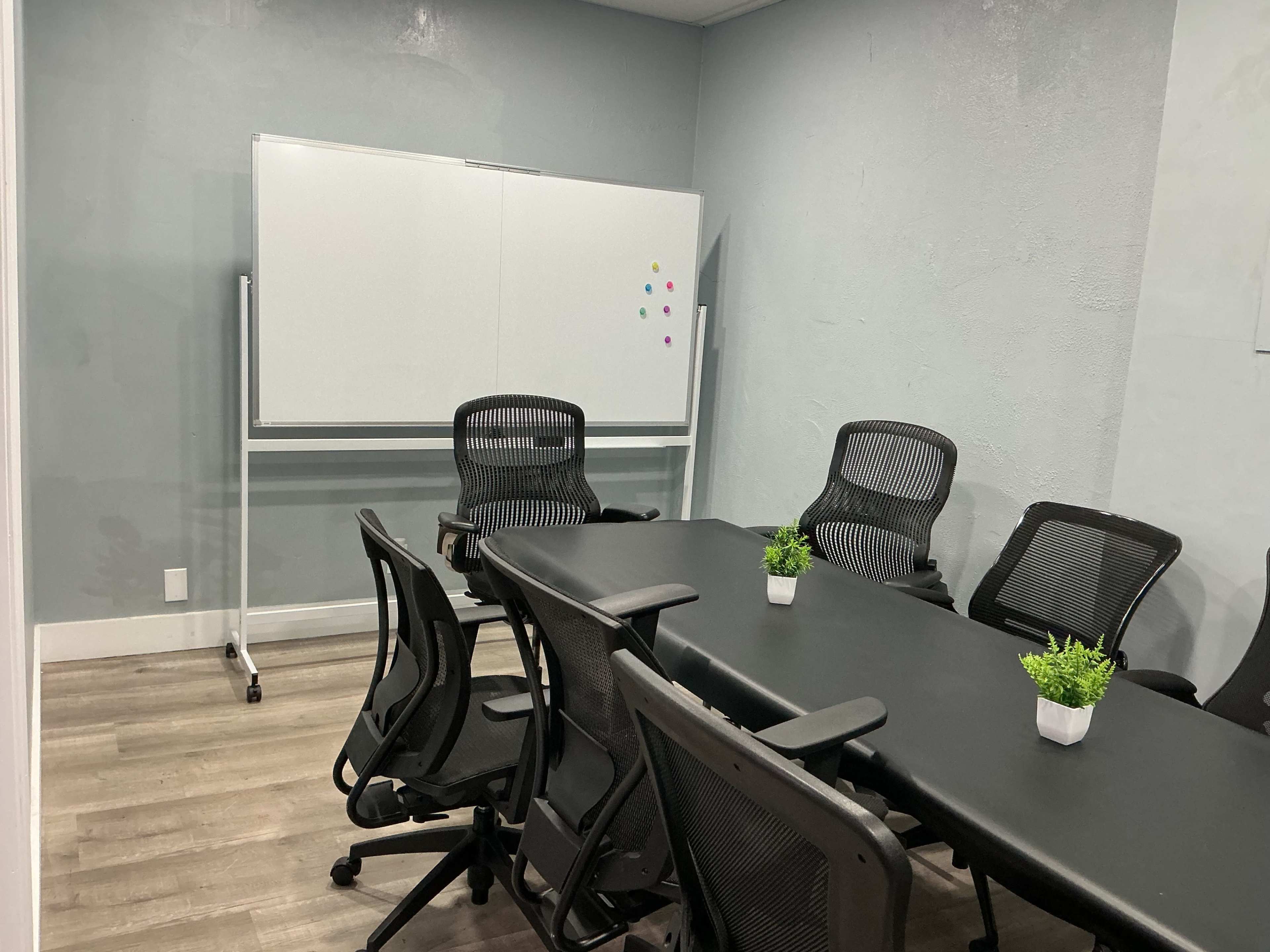 The image shows a small conference room with a long table, black mesh chairs, and a whiteboard on a stand.