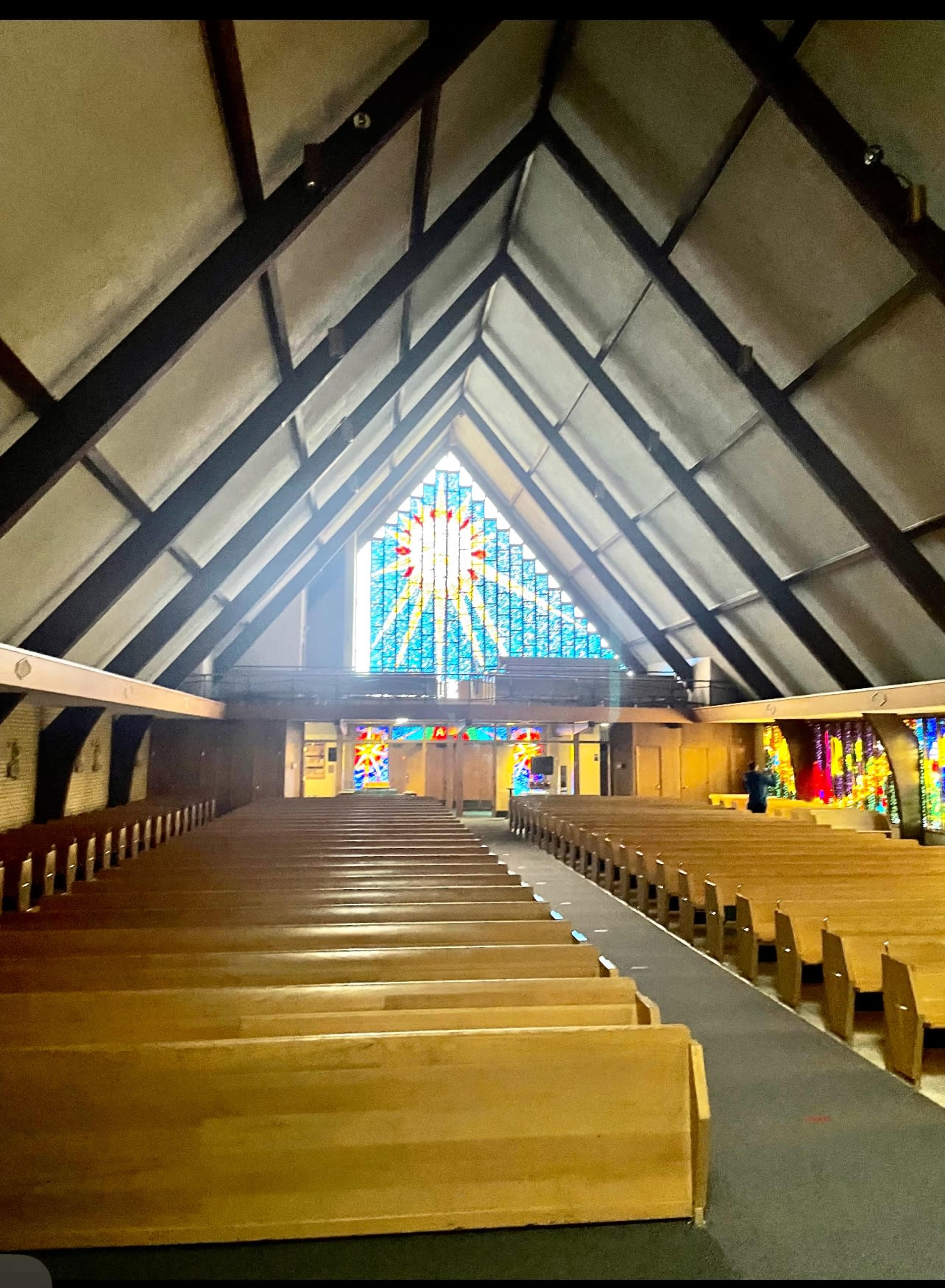 The interior of a church features wooden pews arranged in rows facing a brightly lit stained glass window at the front.