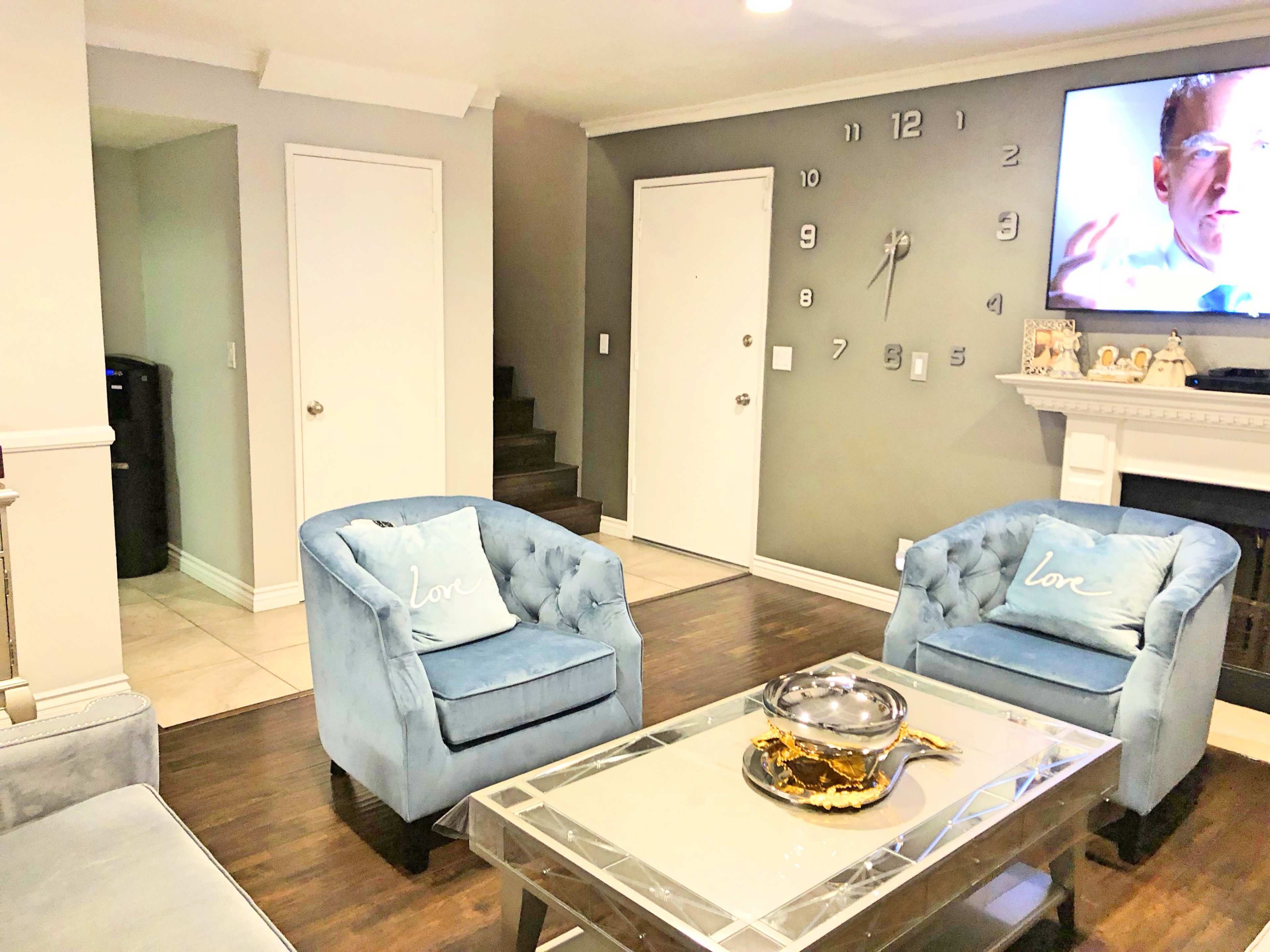 The living room features two blue chairs with decorative pillows, a glass coffee table, and a television mounted on the wall, alongside a staircase and two doors.