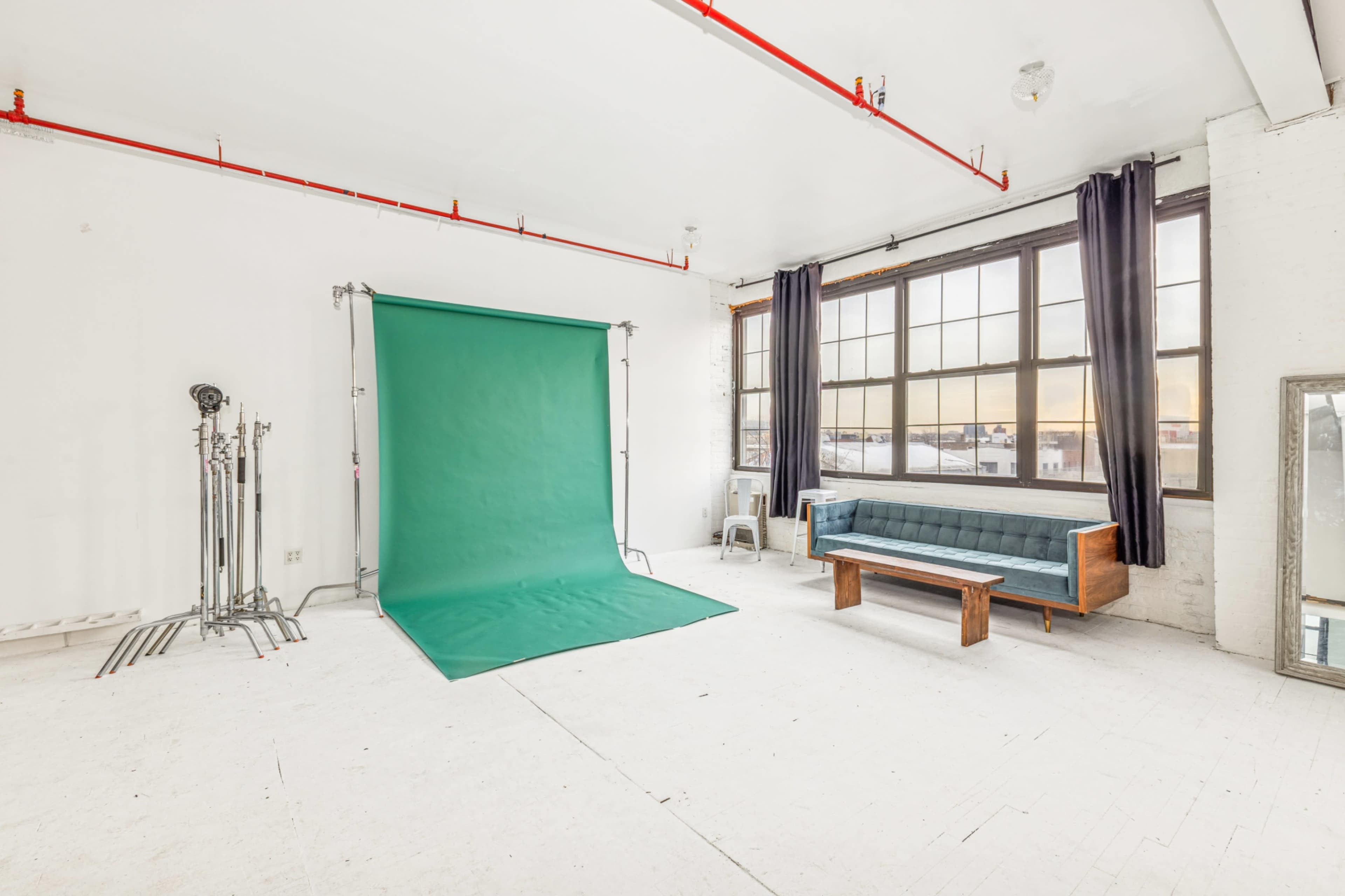 The image shows a spacious, well-lit studio with a green backdrop, a wooden sofa, and large windows.