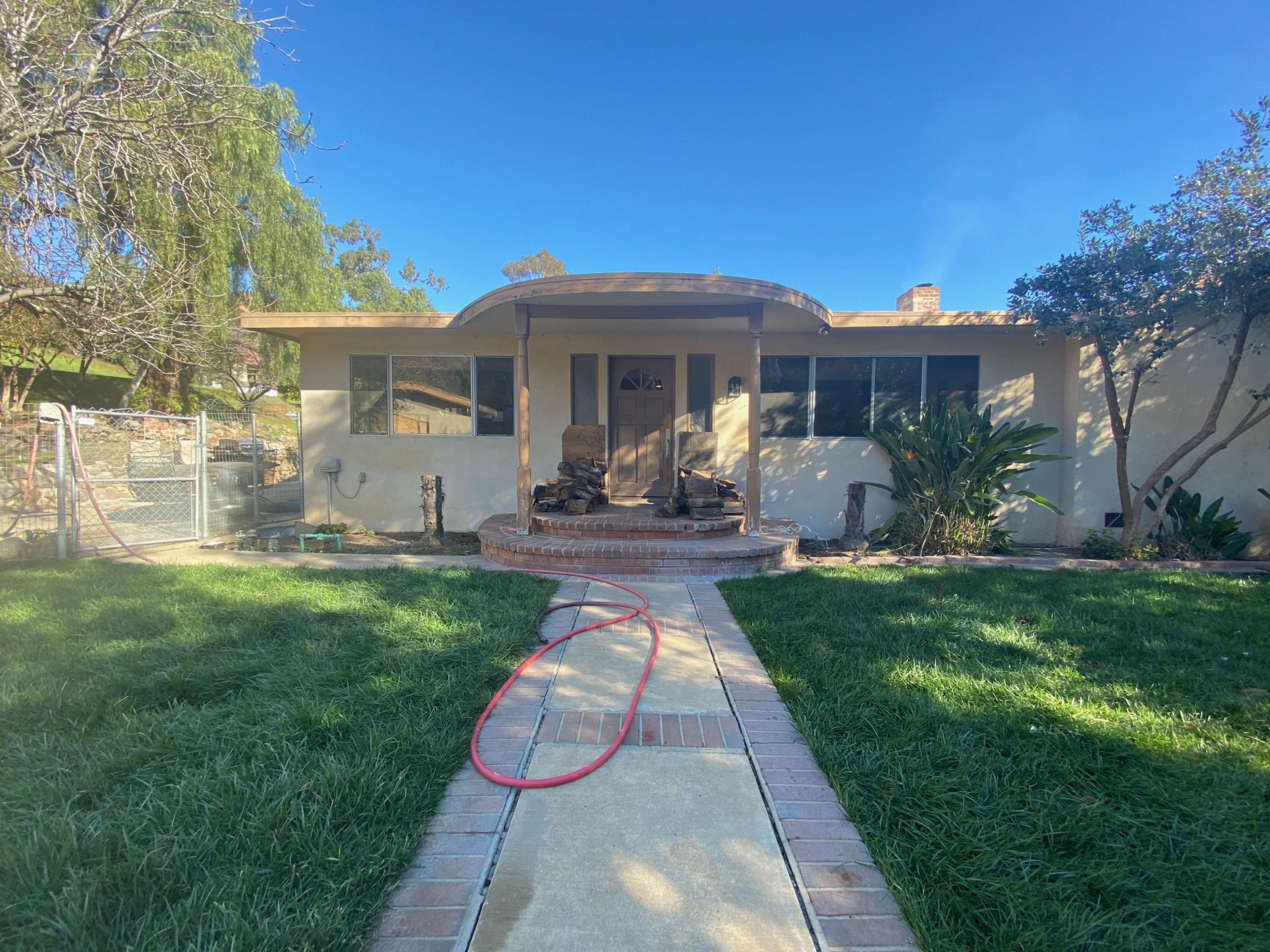 The image shows a single-story house with a front porch, a concrete walkway leading to the entrance, and a yard with grass and shrubs.