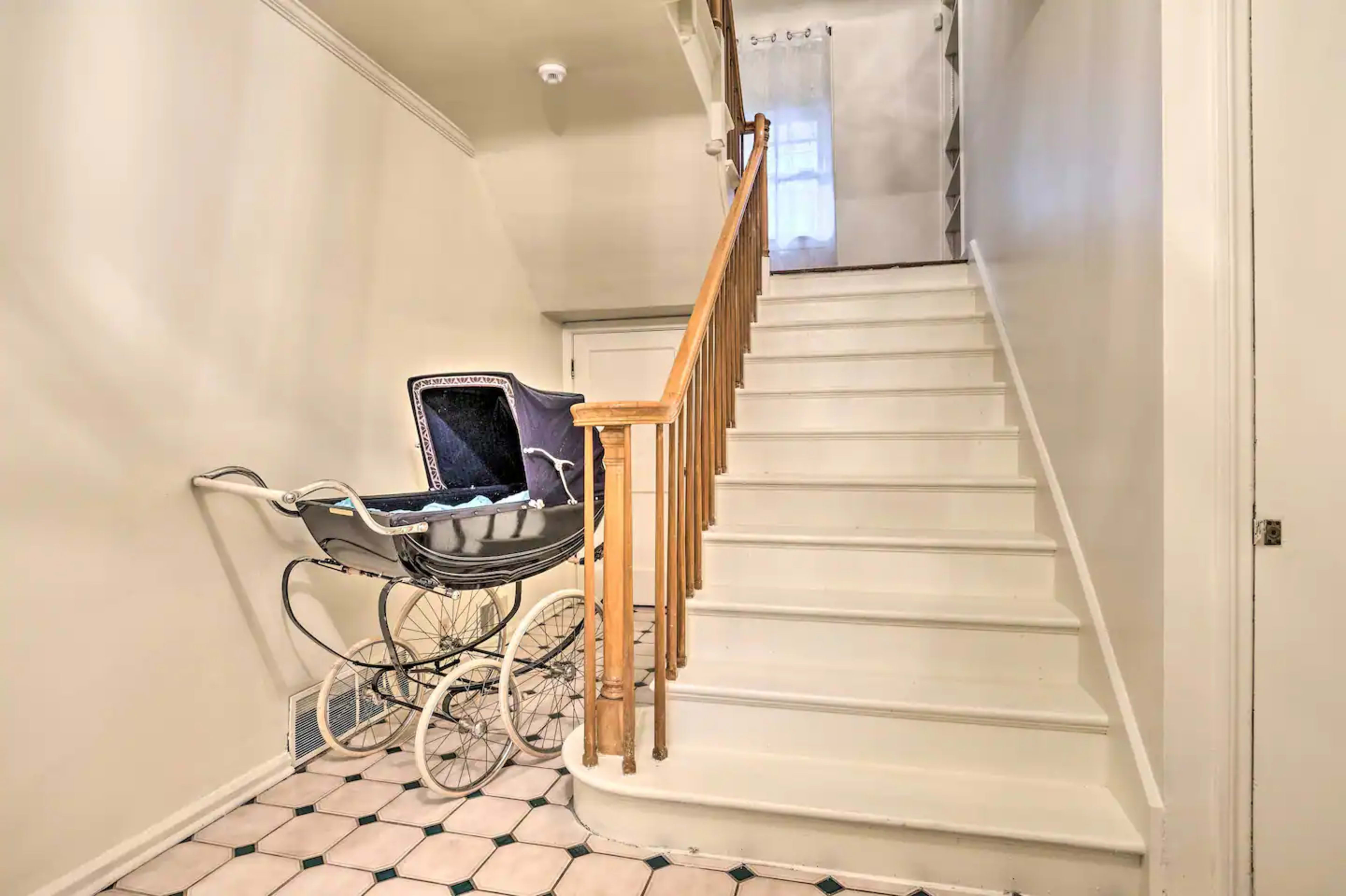 A vintage baby carriage is positioned next to a staircase in a well-lit hallway with tiled flooring.