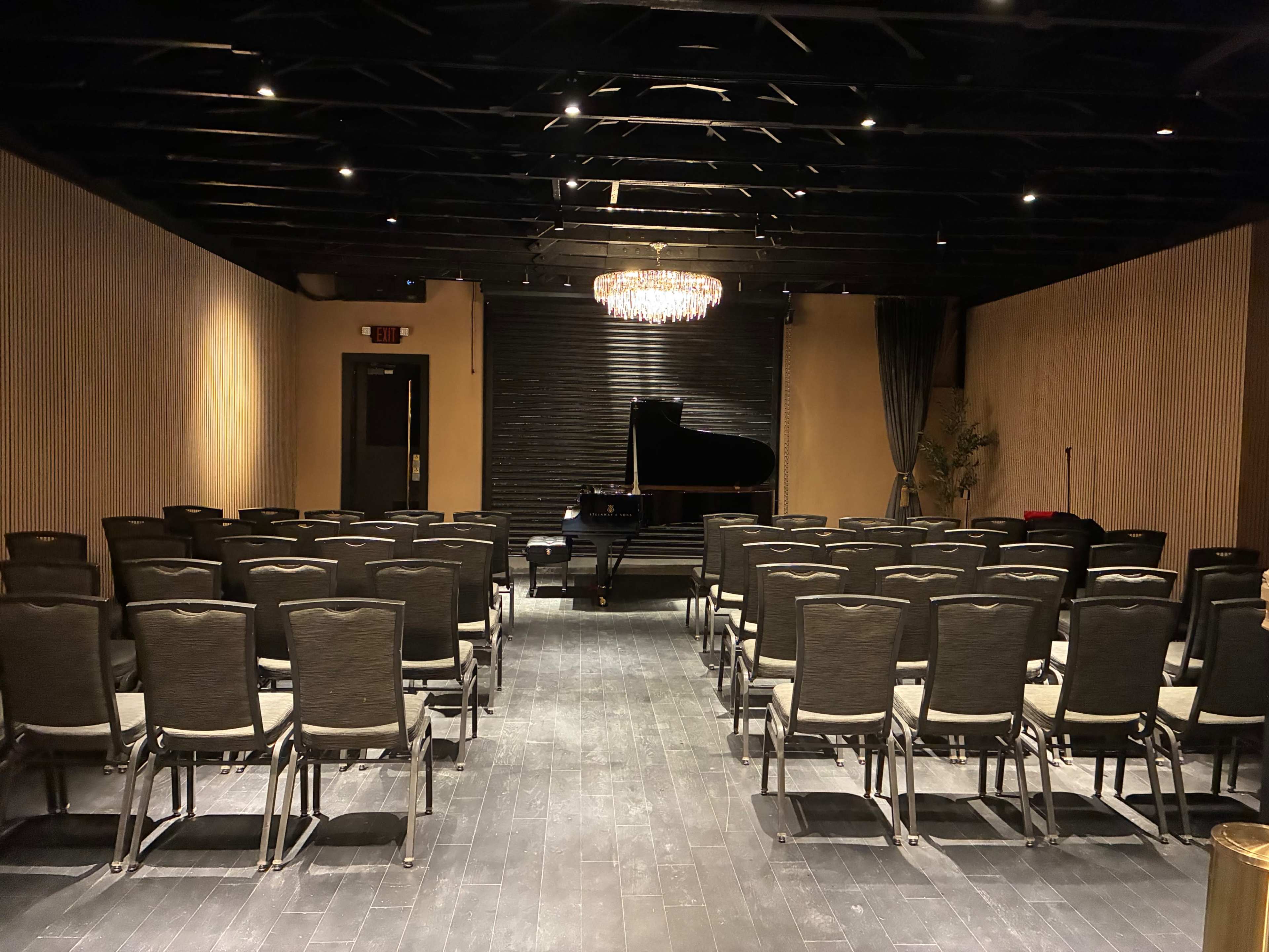 The image shows a dimly lit indoor venue with rows of chairs facing a grand piano and a chandelier overhead.
