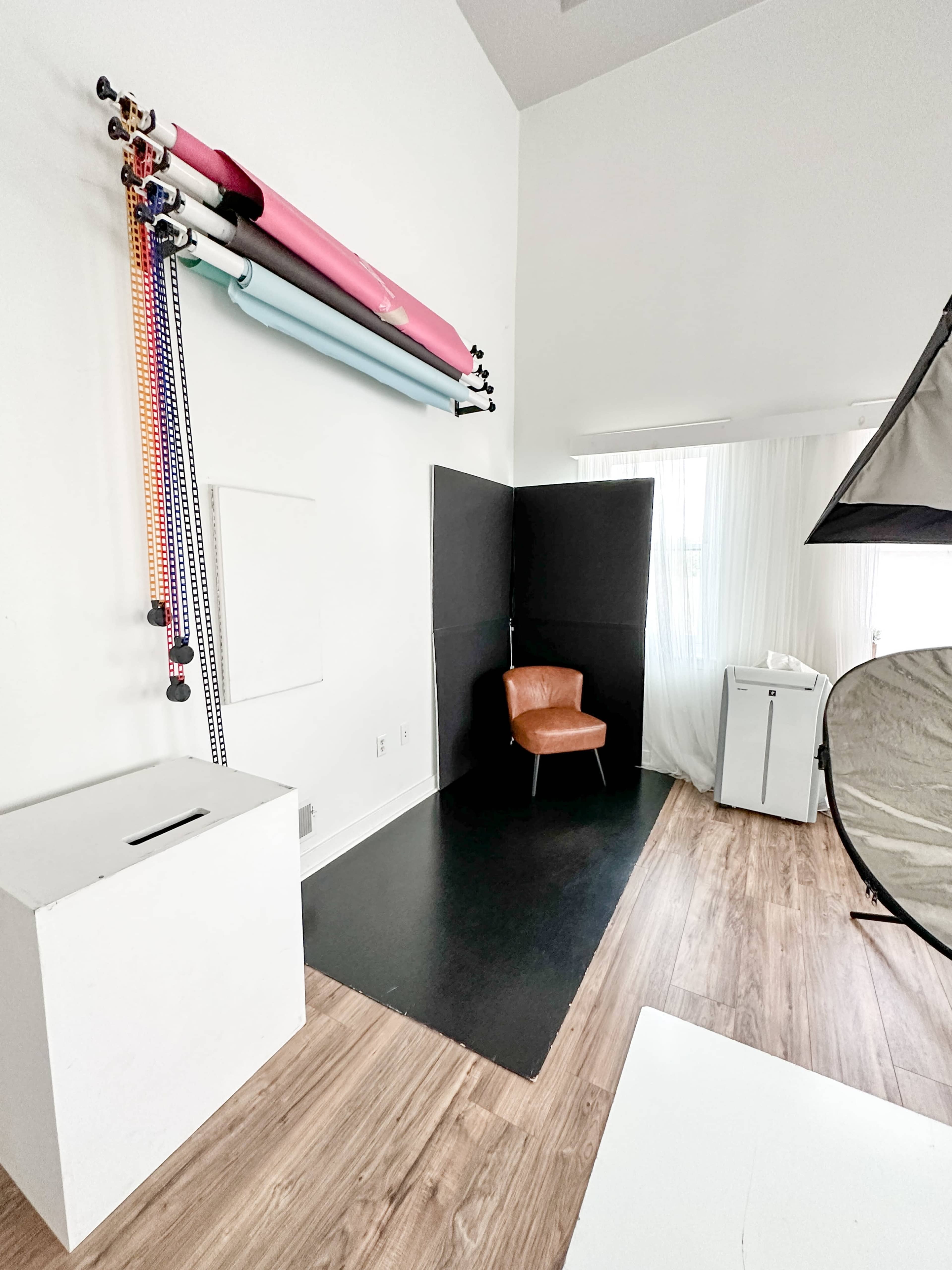A photography studio with a brown chair positioned next to a backdrop stand, rollers of colored paper, and a softbox.
