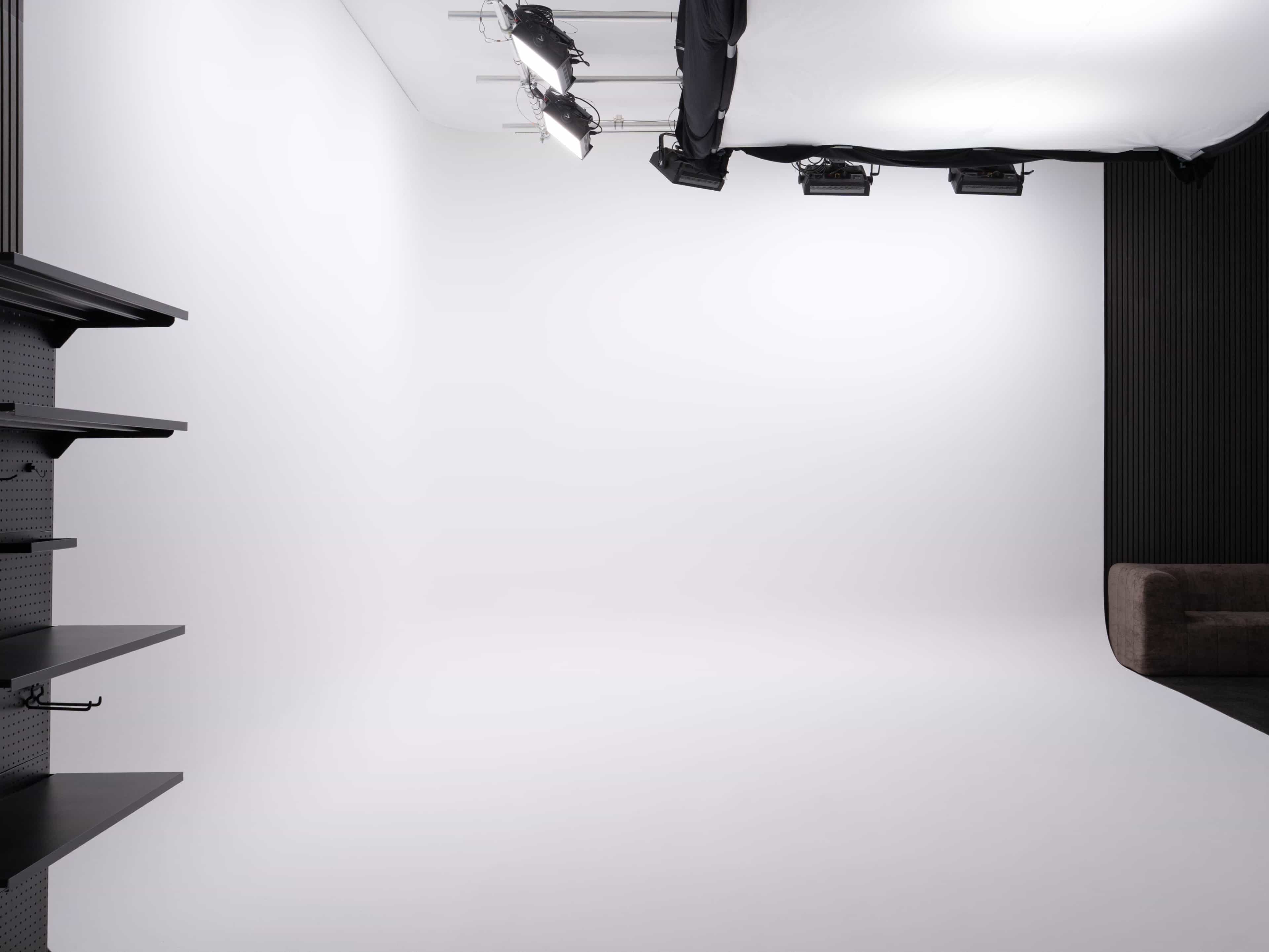 The image shows a minimalistic studio space with a white backdrop, ceiling lights, and a dark sofa along the edge.