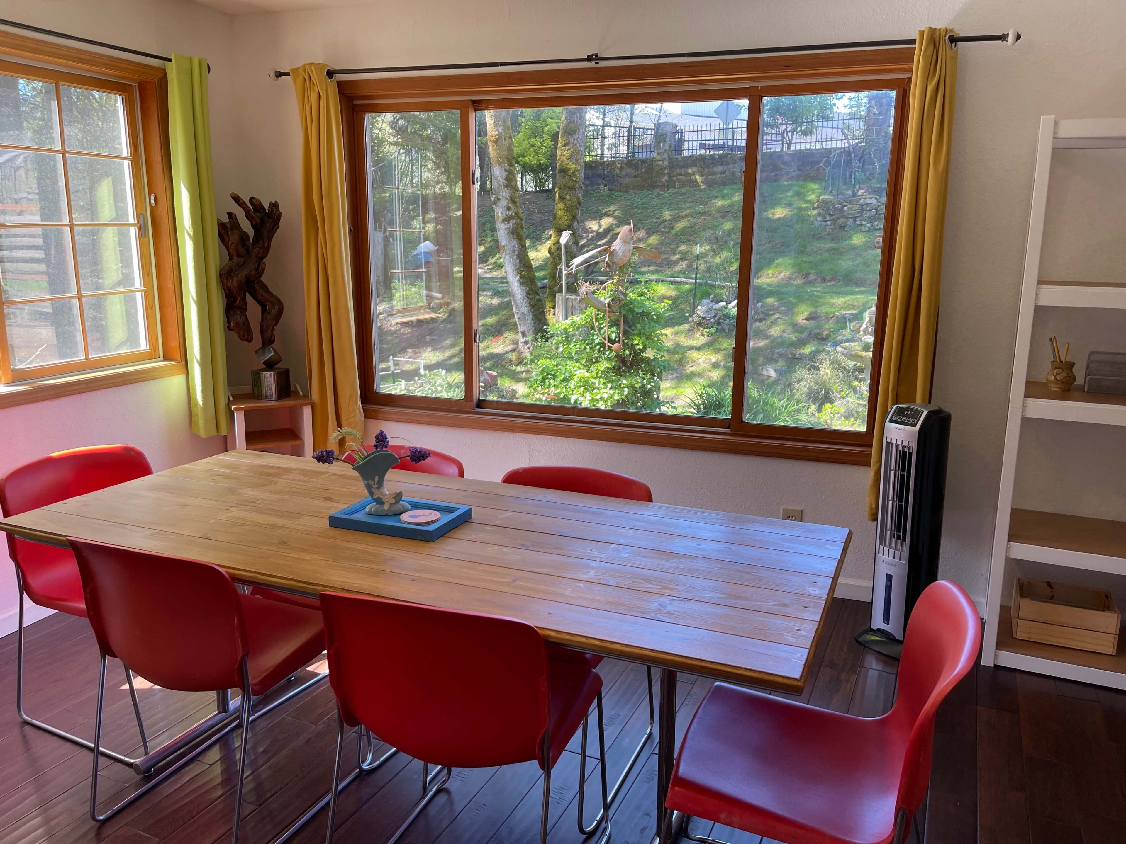 A wooden dining table with red chairs is positioned by a large window overlooking a green yard with trees.