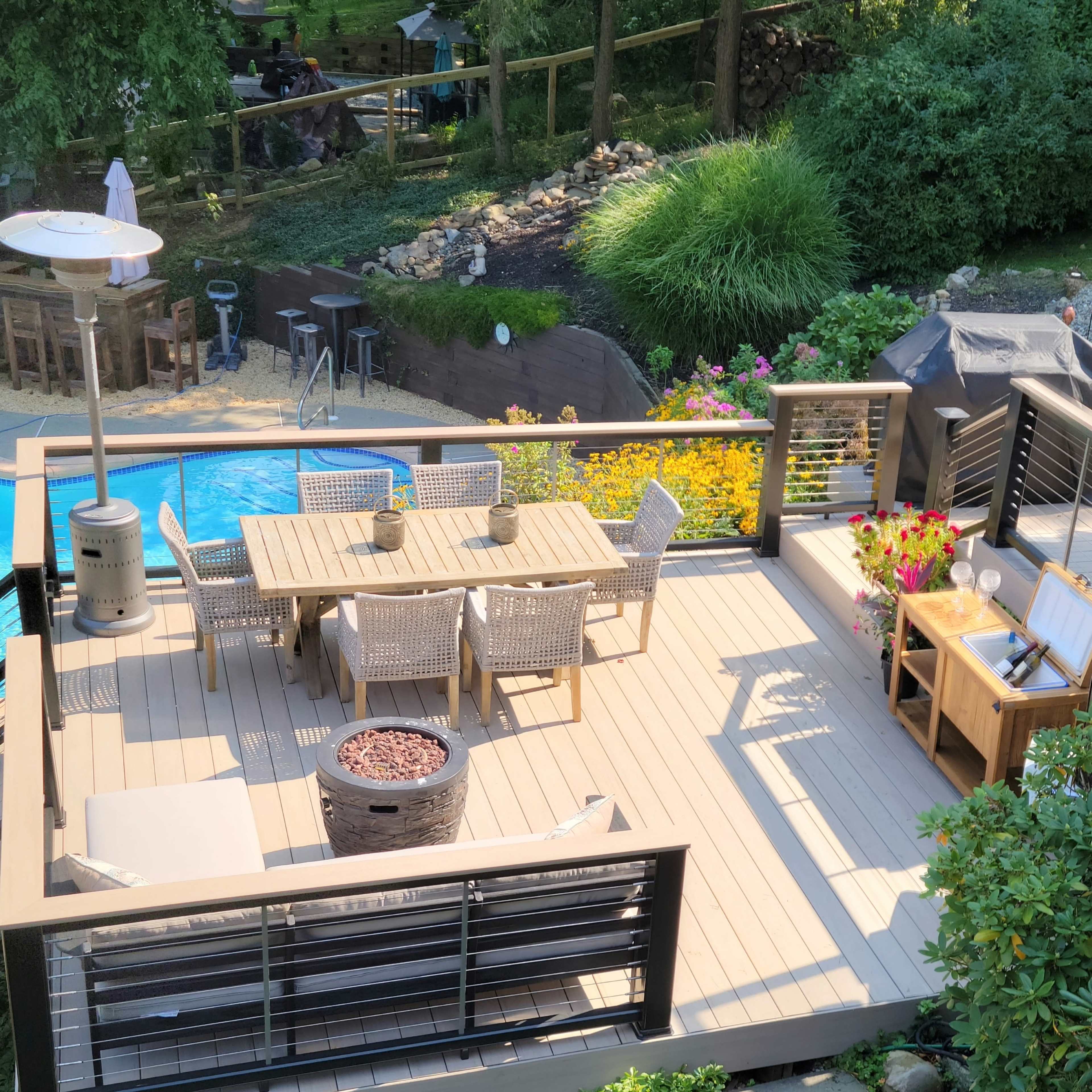 A wooden deck outfitted with a table and chairs, a fire pit, and a view of a garden area with a pool.