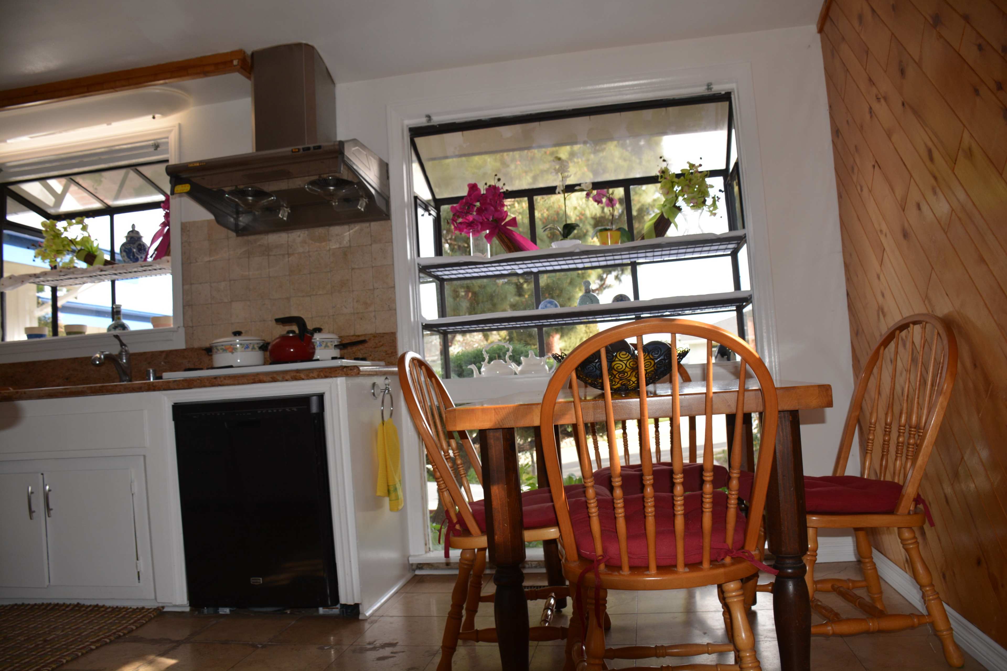 A kitchen with a wooden dining table and chairs, a stove and sink area, and large windows displaying potted plants.